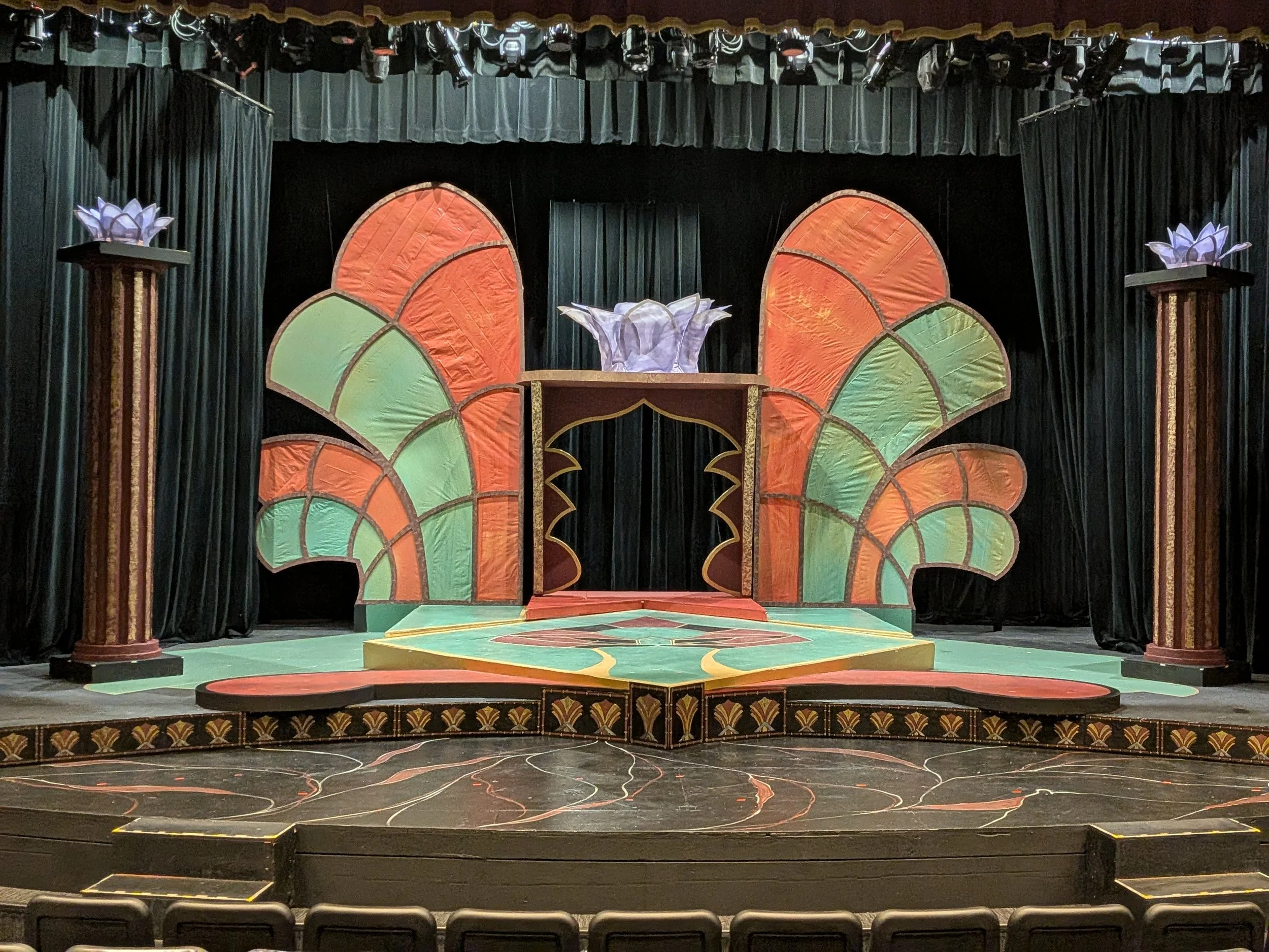 Theater stage decorated with colorful, stylized lotus flower motifs, featuring large green and orange shapes, with dark curtains in the background and lotus-shaped props on tall pillars on each side.
