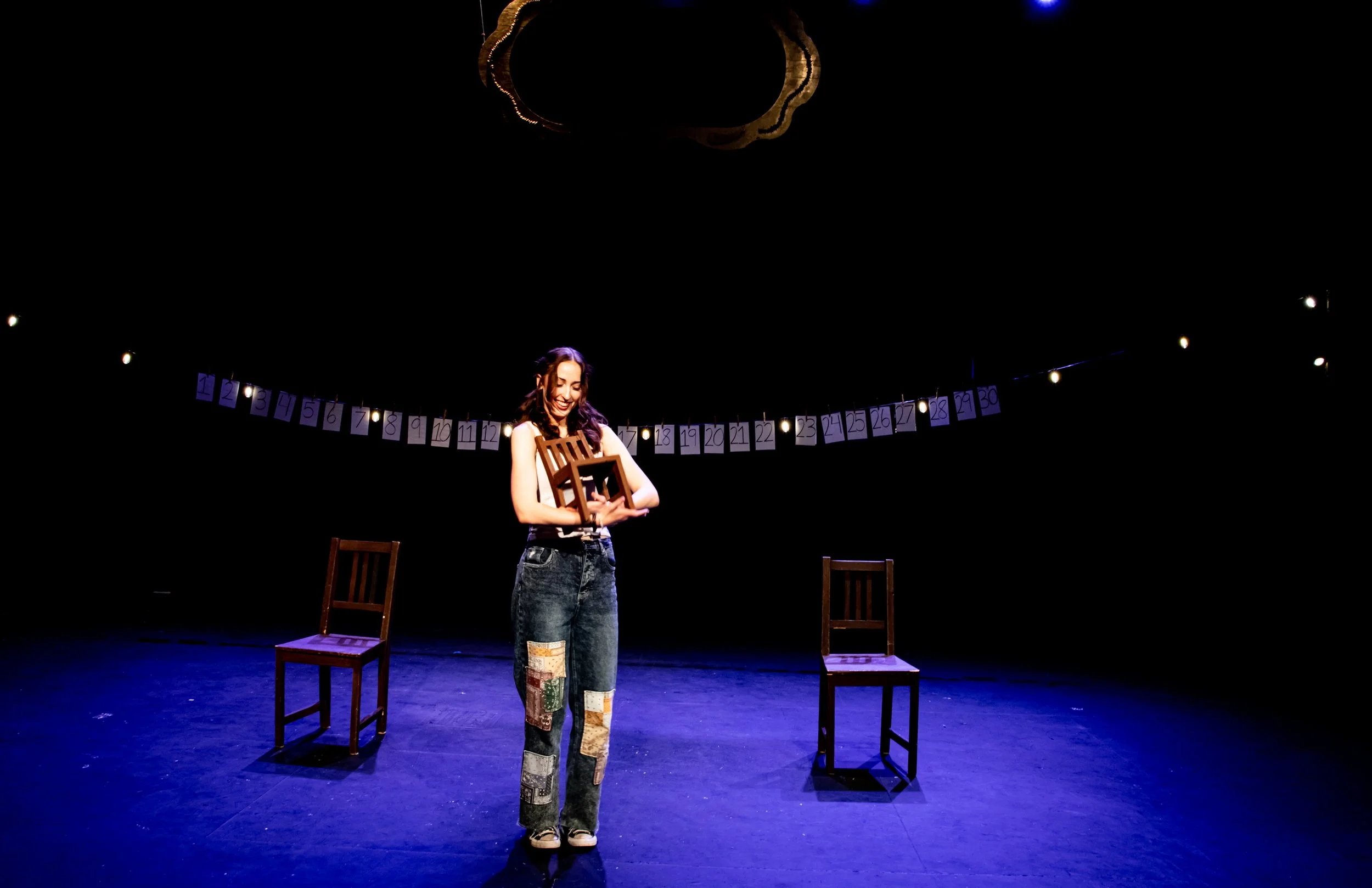 A woman standing on a stage holding a wooden chair, with two additional chairs on either side, and a string of numbered cards hanging behind her illuminated by spotlights.