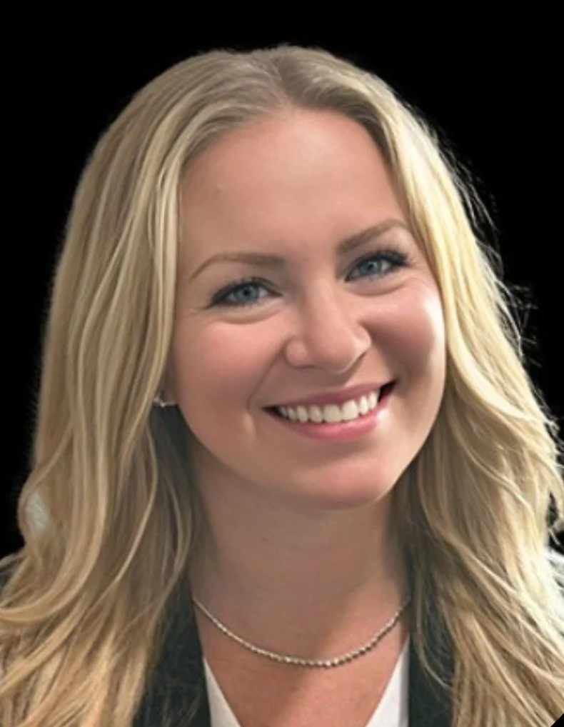 Close-up of a smiling blonde woman with blue eyes wearing a pearl necklace and a dark blazer, with a black background.