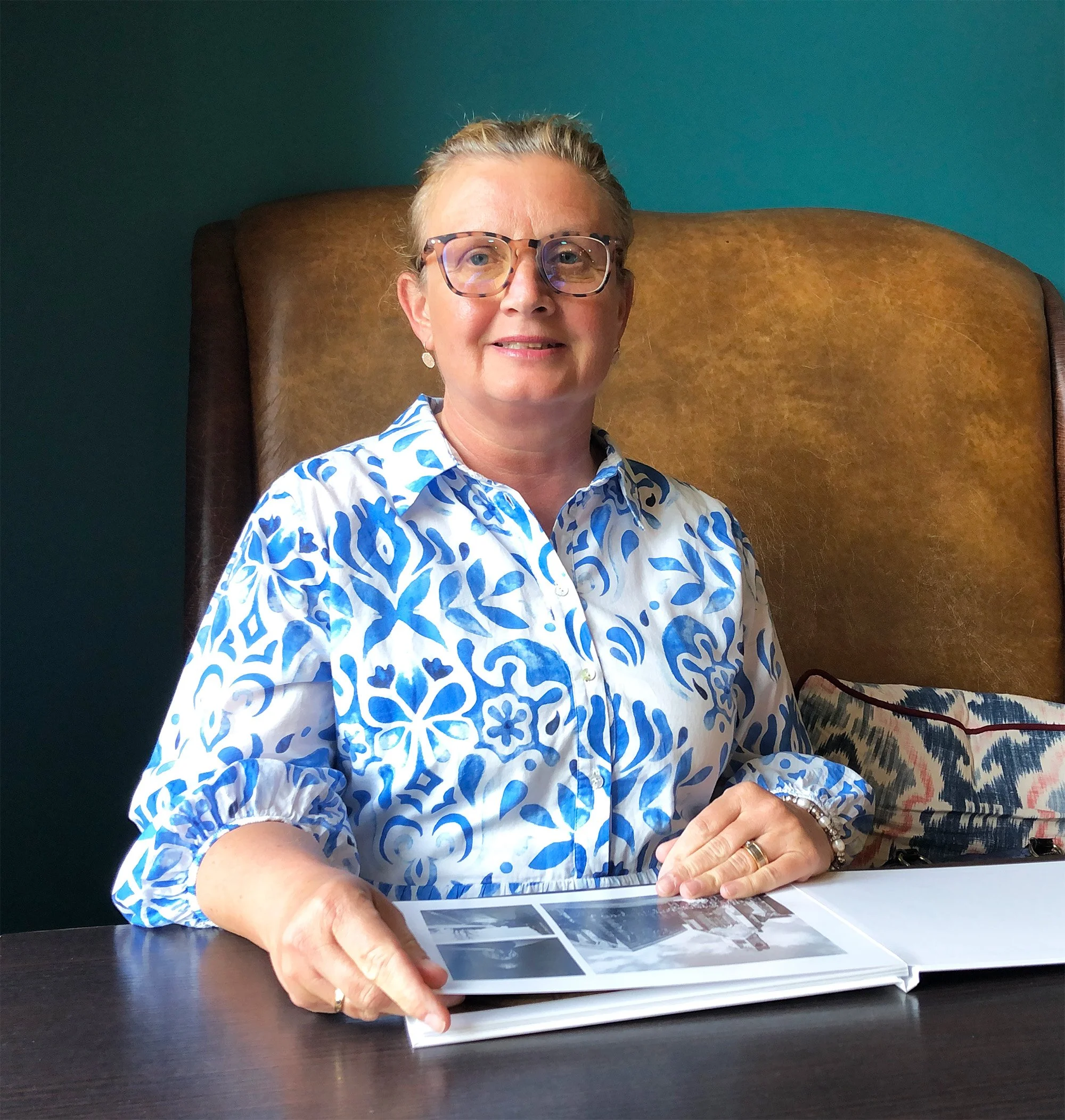 Eine ältere Frau mit kurzen, blonden Haaren und Brille sitzt an einem Tisch und schaut in die Kamera. Sie trägt eine weiße Bluse mit blauen Mustern und liest in einem Fotoalbum. Der Hintergrund ist eine dunkelgrüne Wand.