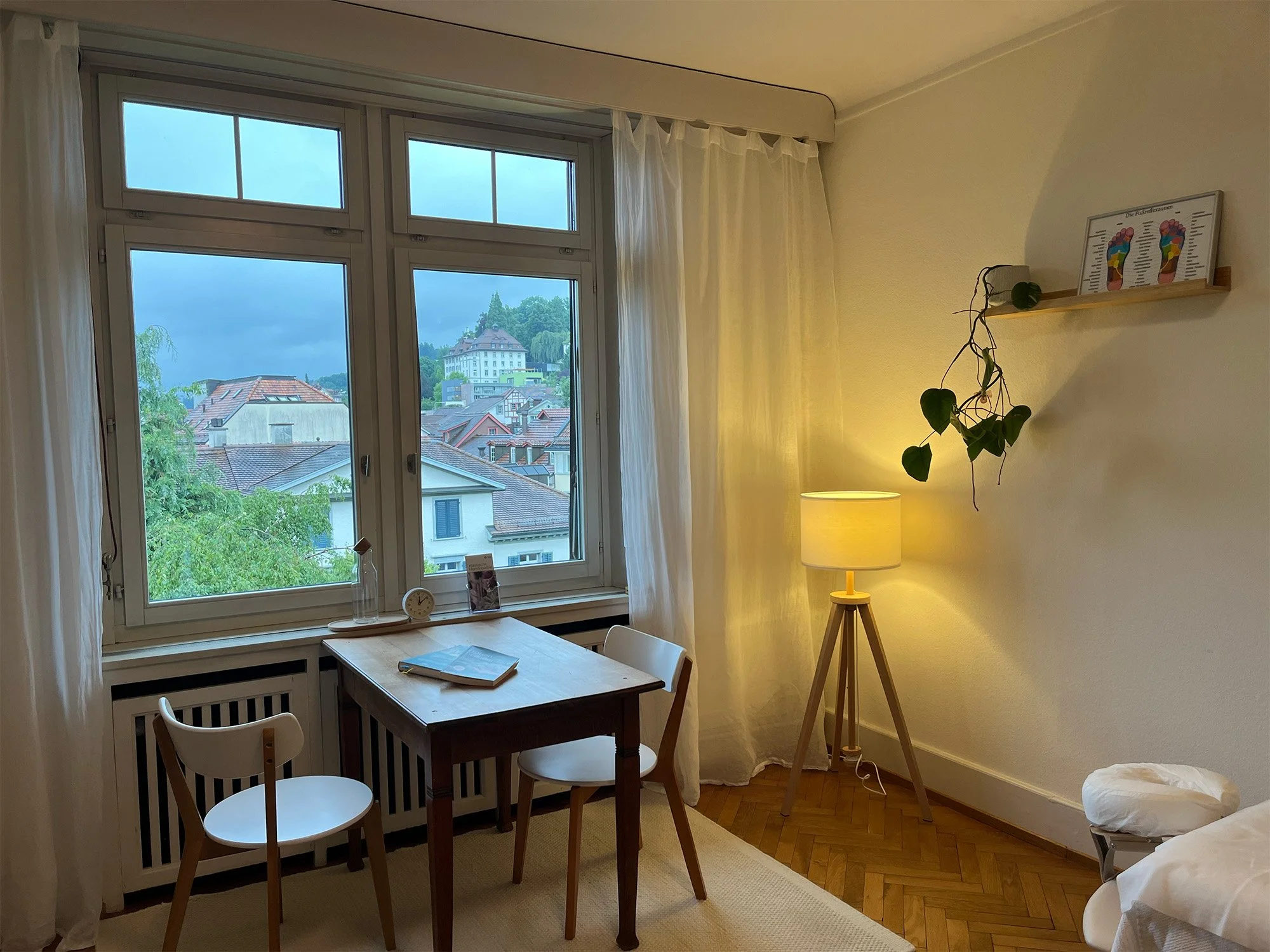 Ein Zimmer mit großem Fenster, einem kleinen Holztisch, zwei weißen Stühlen, einem Stehlampe, einem Wandbild mit Fußreflexzonen, einem Pflanzenkaktus und einem Bett.