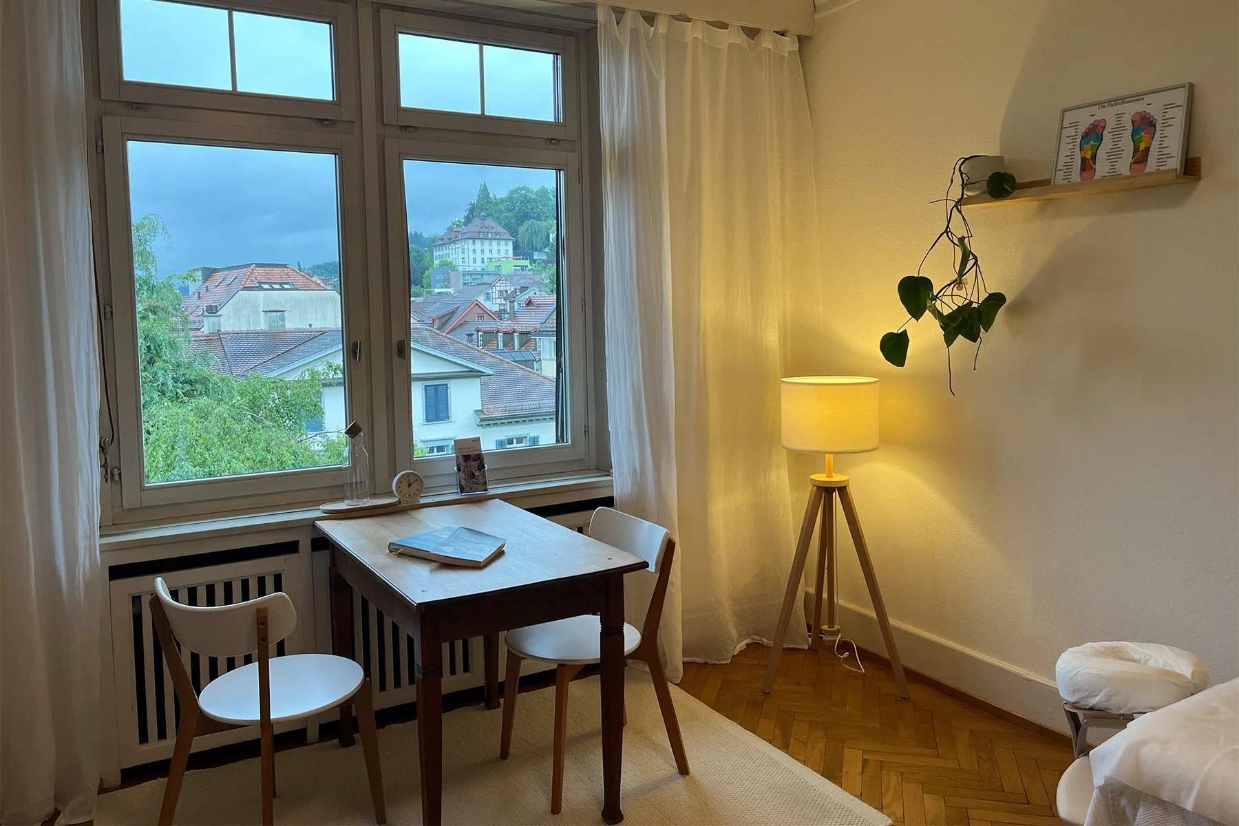 Ein Zimmer mit einem Fenster, einem kleinen Holztisch, zwei Stühlen, einer Stehlampe, einer Wandregal mit Fußreflexzonen-Reflexzonen-Massage-Tafel und einer weißen Bettwäsche.