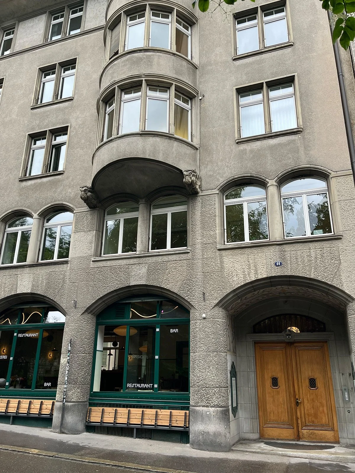 Ein mehrstöckiges Gebäude mit Steingestaltung, großen Fenstern und einem Holzportal. Im Erdgeschoss sind ein Restaurant, eine Bar und eine Bäckerei zu sehen.