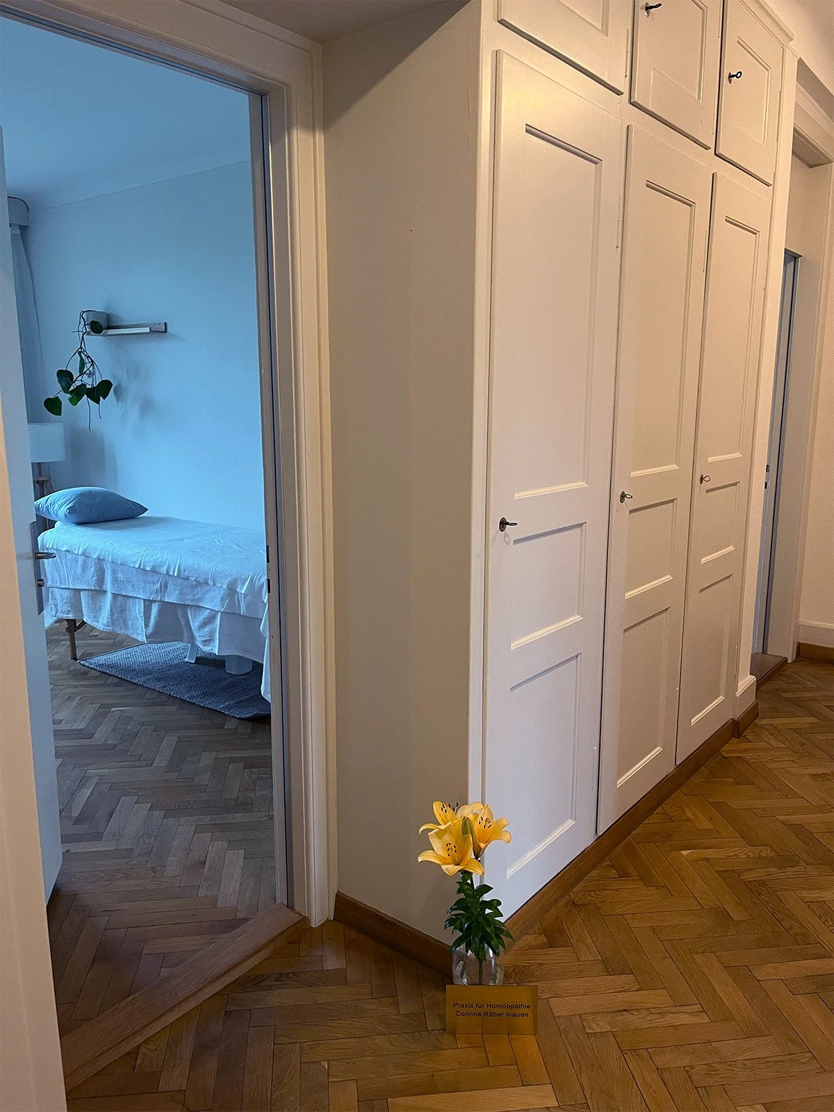 A vase mit gelben Lilien auf dem Boden neben einem Schild, das auf eine Homöopathie-Praxis verweist, steht vor weißen Schränken in einem Flur.