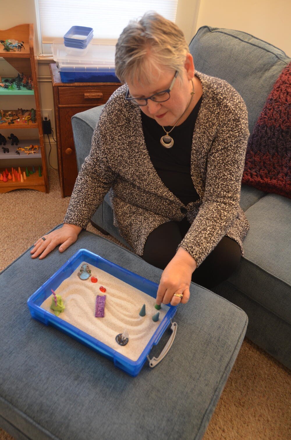 Heal Grow Go Wellness therapist Helen Svoboda-Barber sitting on a blue couch with a sand tray.