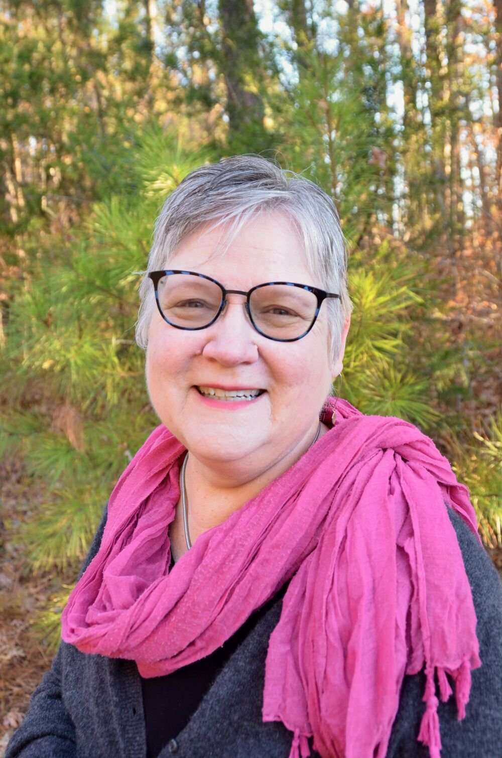 A smiling woman with short gray hair, glasses, wearing a pink scarf and dark top, standing outdoors with trees in the background.