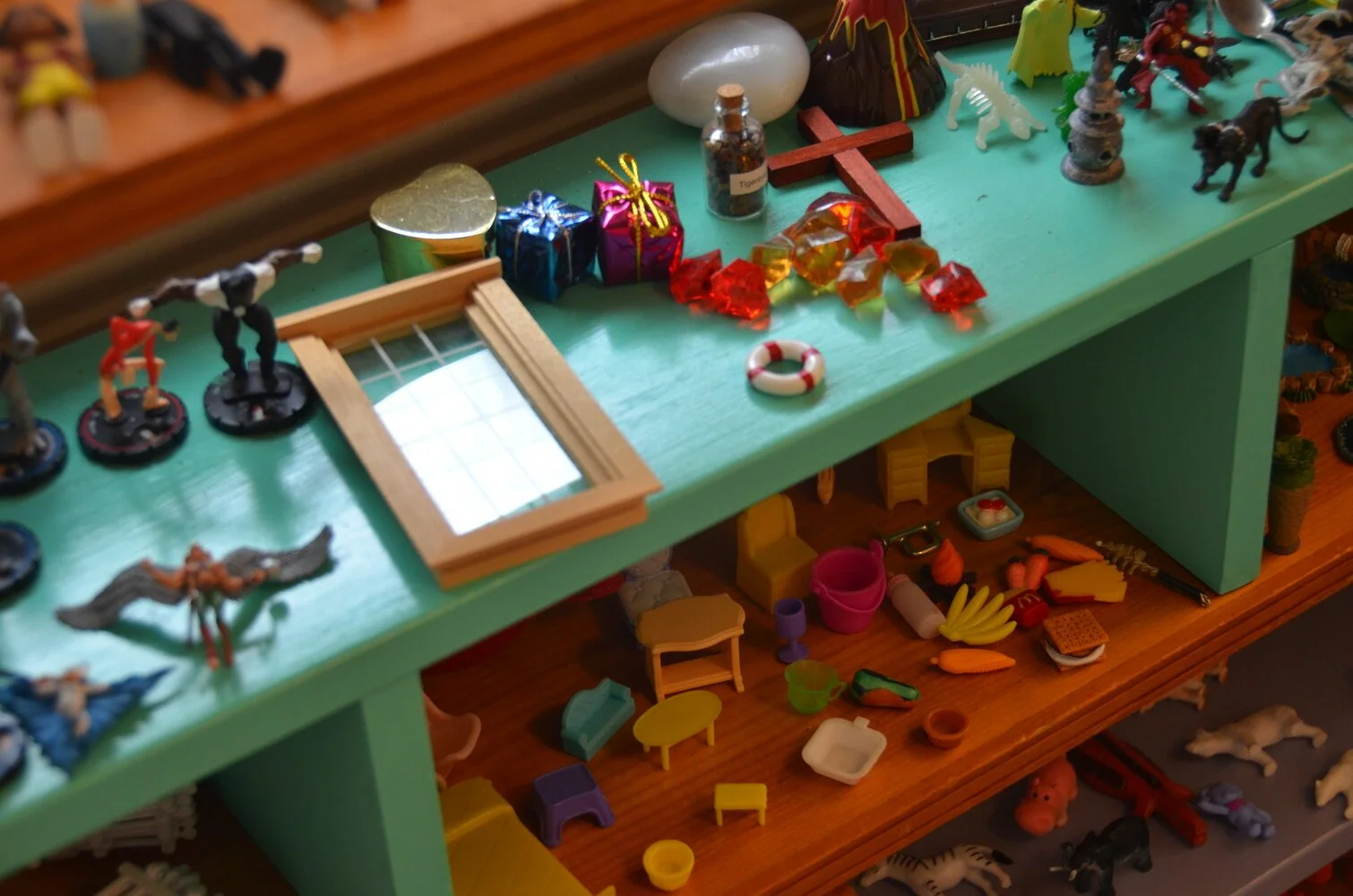 A green display table with small toys and figurines, including a miniature life preserver, wrapped presents, a small cross, a glass bottle labeled 'Tigers,' and various tiny furniture pieces and accessories.