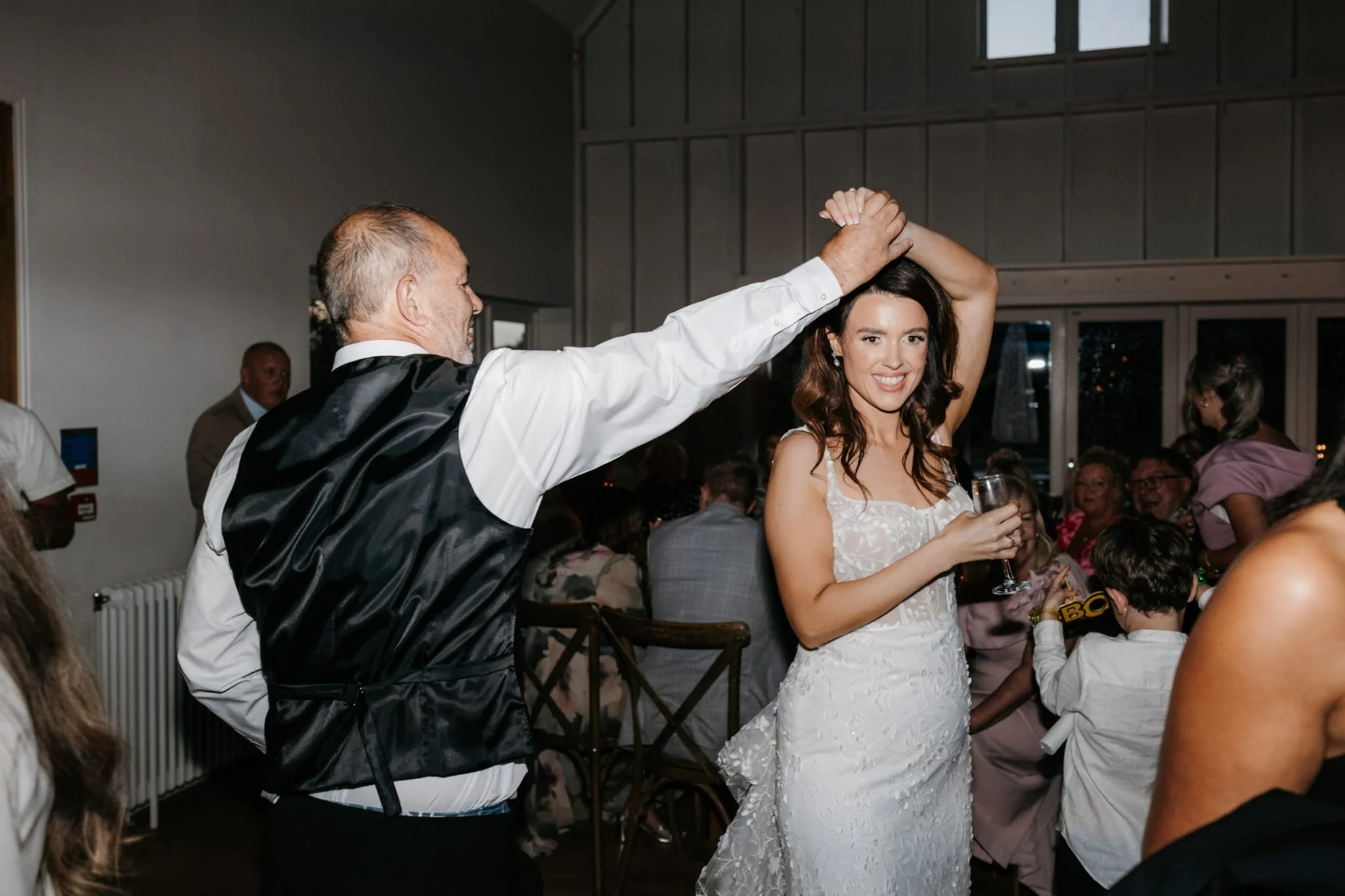 An older man in a black vest and white shirt dancing with a young woman in a white wedding dress at a wedding reception.