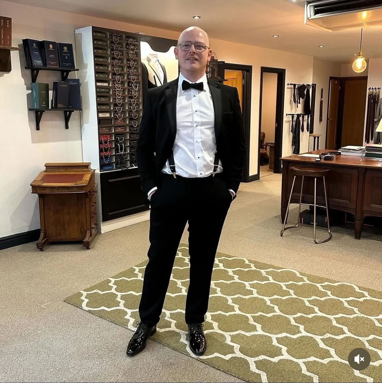 A man wearing a tuxedo with a white shirt, black bowtie, and black dress shoes standing in an office or showroom space.