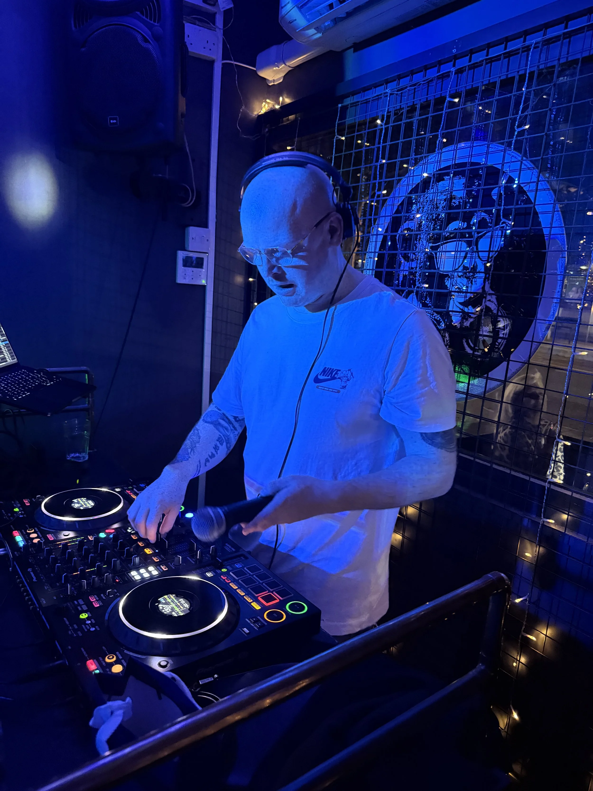 DJ with tattoos wearing glasses and headphones, mixing music at a nightclub with blue lighting, behind a wire fence, and a neon clock in the background.