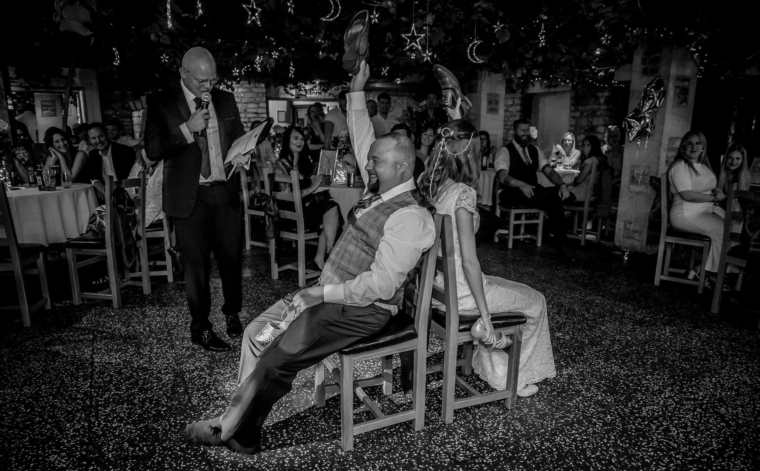 A black-and-white photo of a wedding reception with a man and woman sitting back-to-back on chairs, playing a game. The man is laughing and has his arm raised. Guests are seated at tables in the background, with decorations like hanging stars and moons.