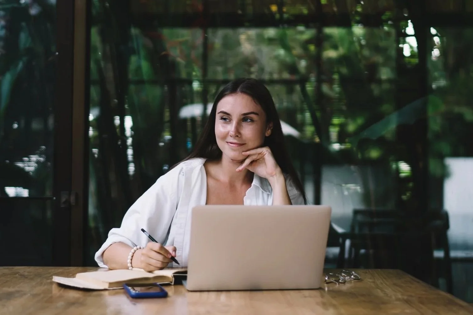 A woman sitting at a wooden table with a laptop, notebook, and smartphone in front of her, looking thoughtfully to the side inside a modern, glass-walled building surrounded by greenery.
