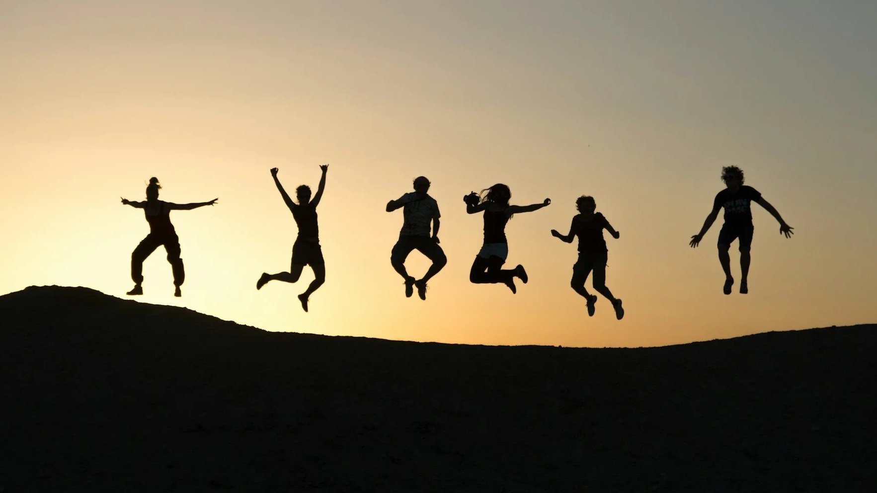 Silhouettes of six children jumping in the air on a hill against a sunset sky.