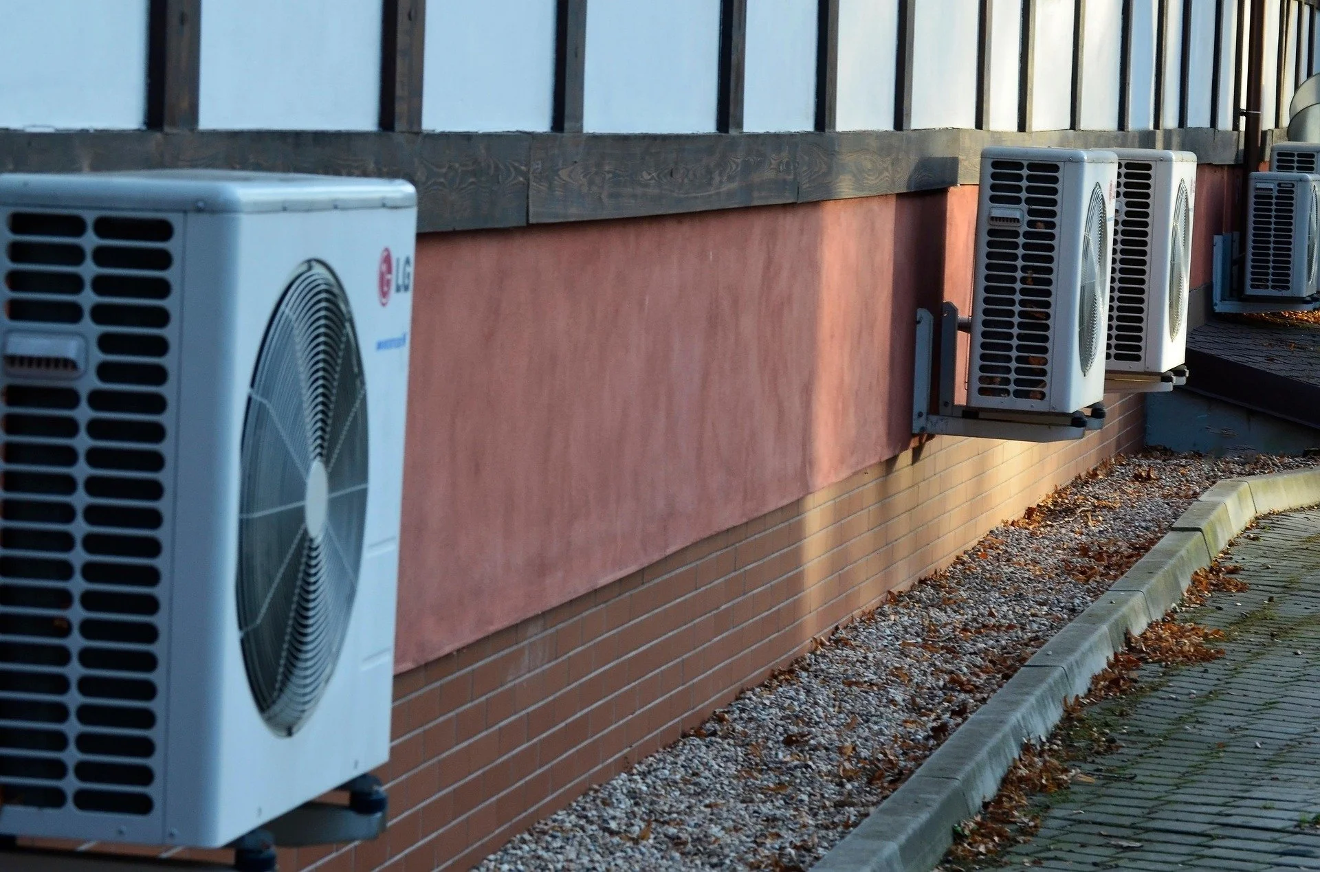 Mehrere Klimaanlagen sind an einer roten und braunen Hauswand installiert, entlang eines Fußweges mit Laub und Kies.