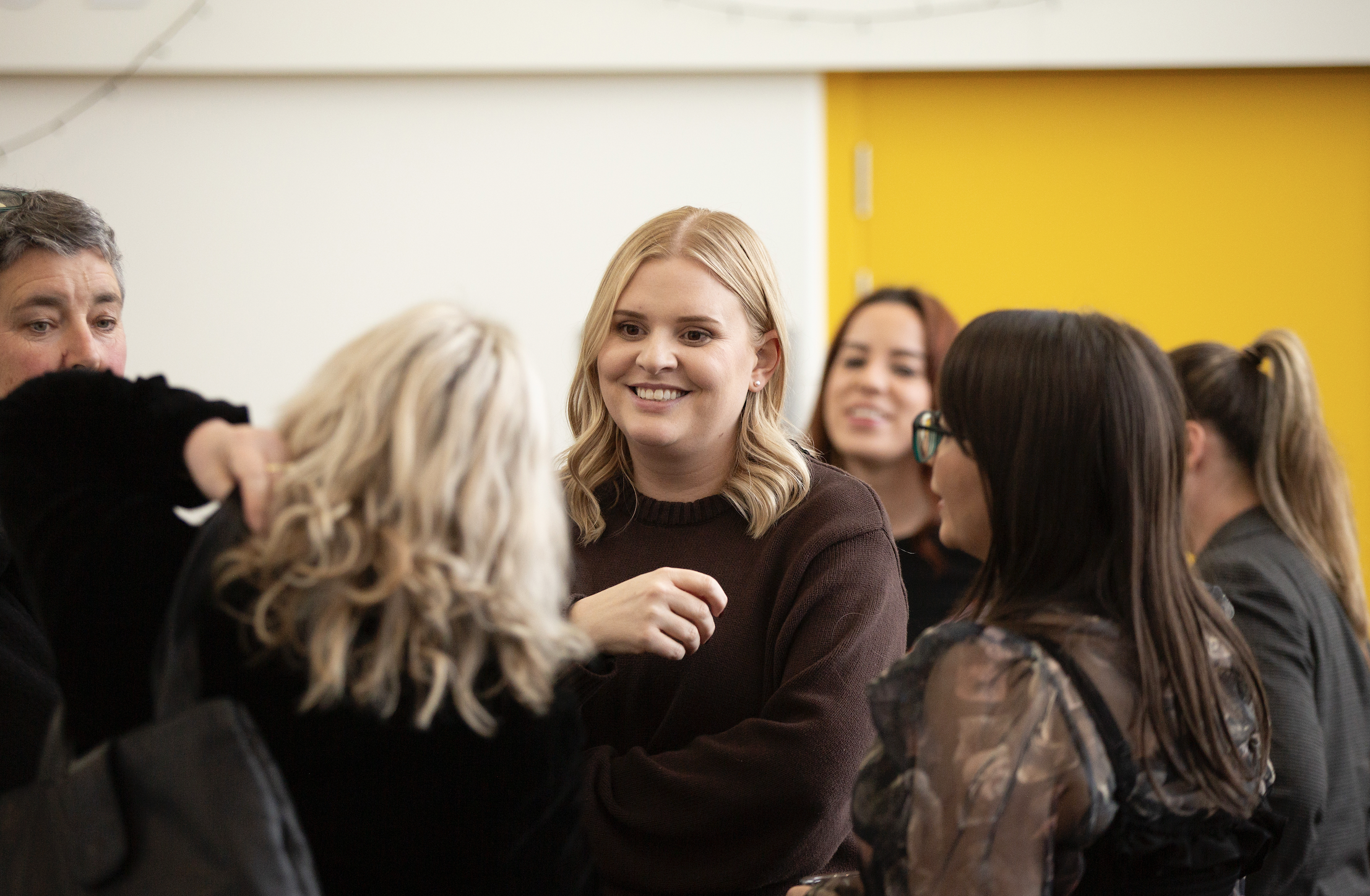 A group of diverse women and men engaged in conversation indoors, smiling and enjoying each other's company.