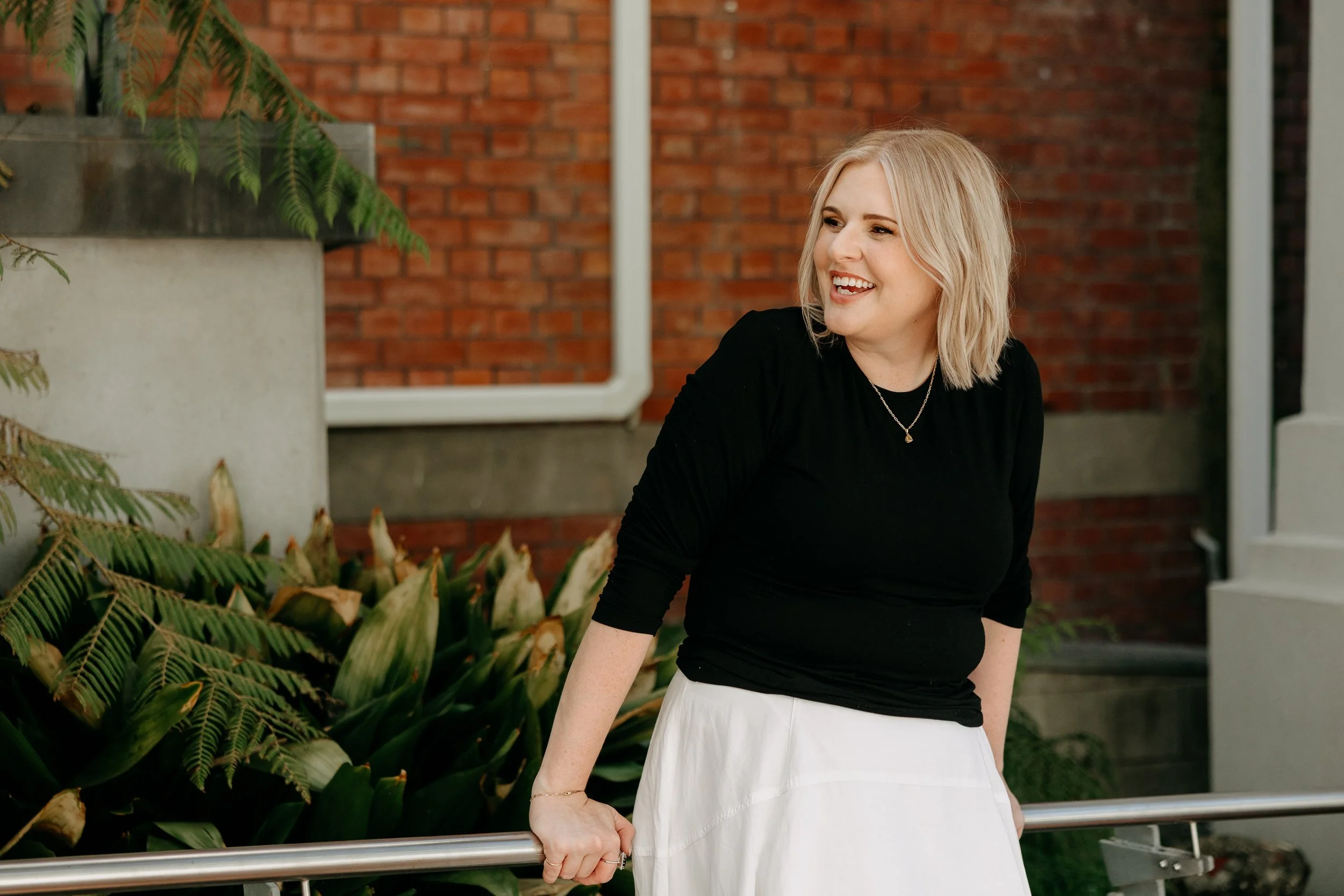A woman with blonde hair and a black top smiling outdoors near plants and a brick wall.