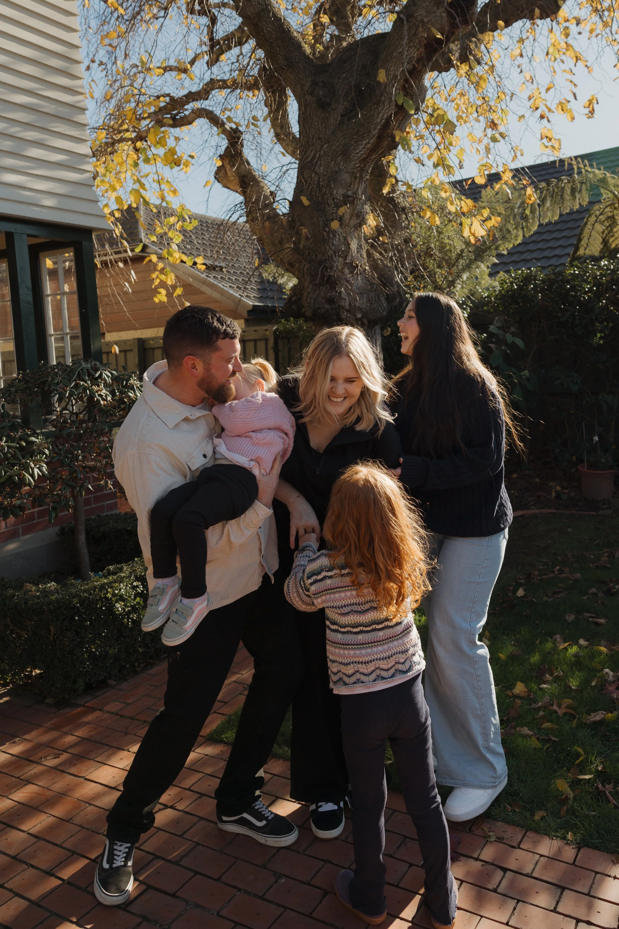 A group of five people, including two children and three adults, smiling and playing together outdoors on a brick patio with a large tree and house in the background during autumn.