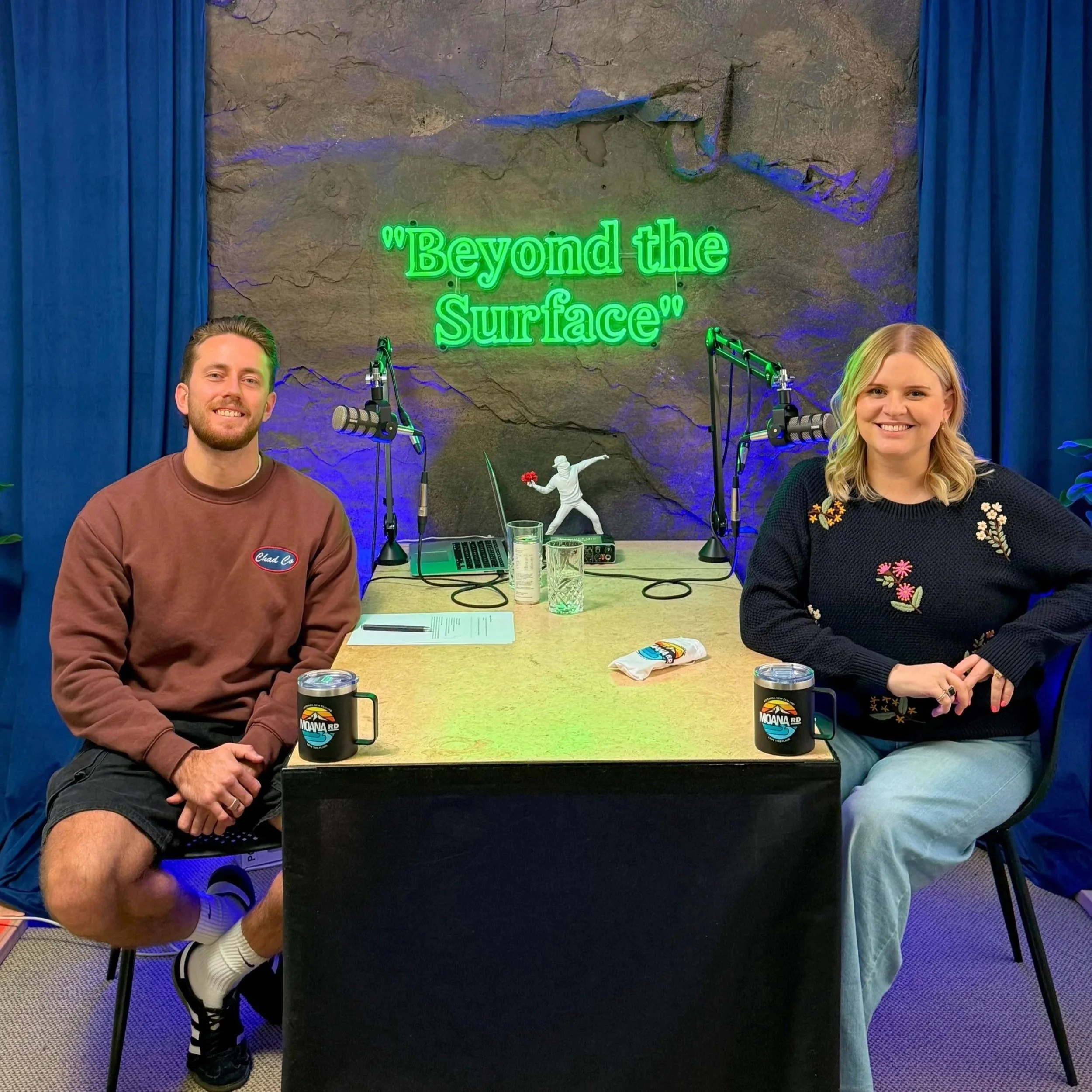 Two people sit at a table with microphones, in a studio with blue curtains and a neon sign that reads 'Beyond the Surface'.