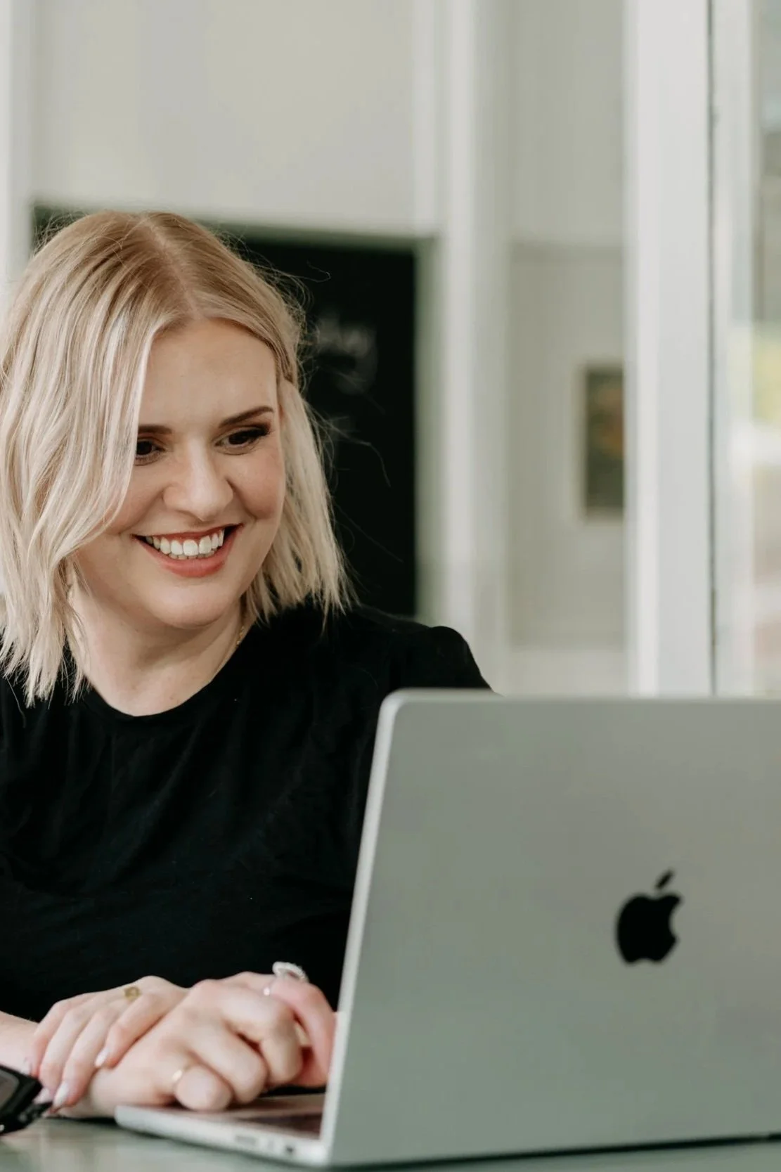A woman (Lizi Guest) with blonde hair smiling and working on a MacBook laptop in a bright room.