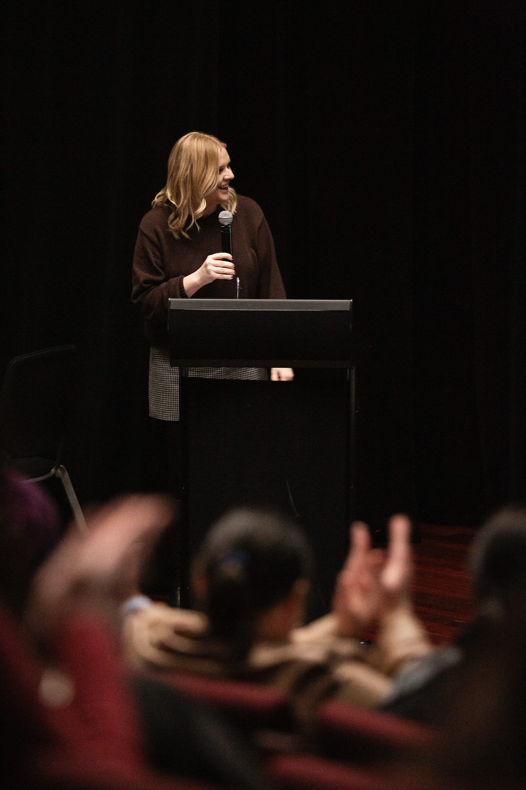 A woman standing at a podium, holding a microphone, smiling and looking to her right while speaking to an audience.