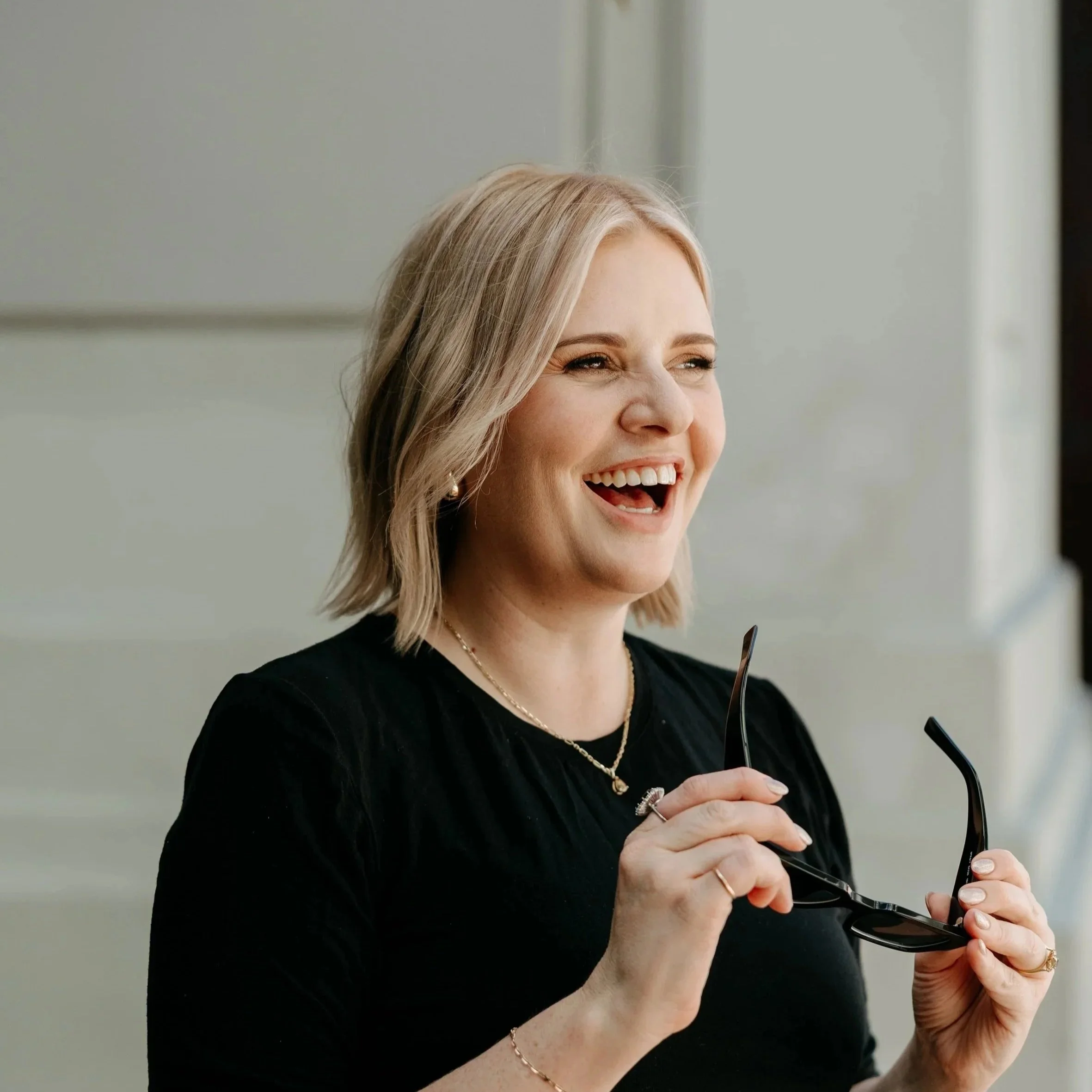 A woman (Lizi Guest) with shoulder-length blonde hair smiling while holding sunglasses.