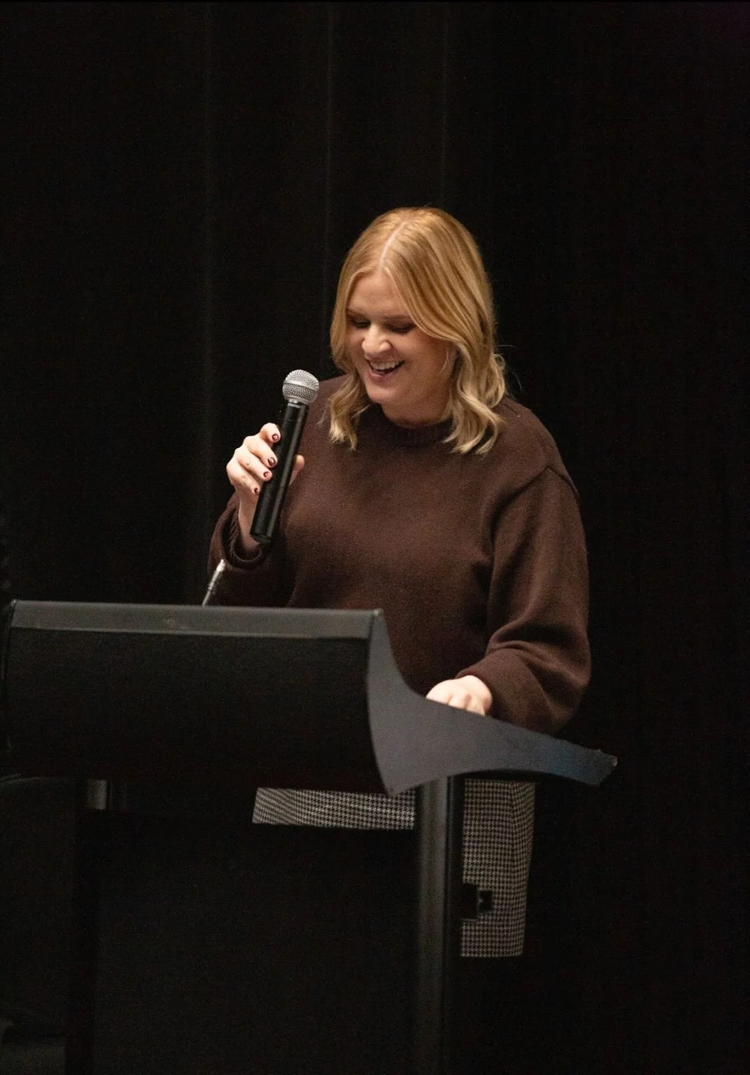A woman with blonde hair holding a microphone in front of a music stand, smiling and looking down, wearing a brown sweater, in a dark room.