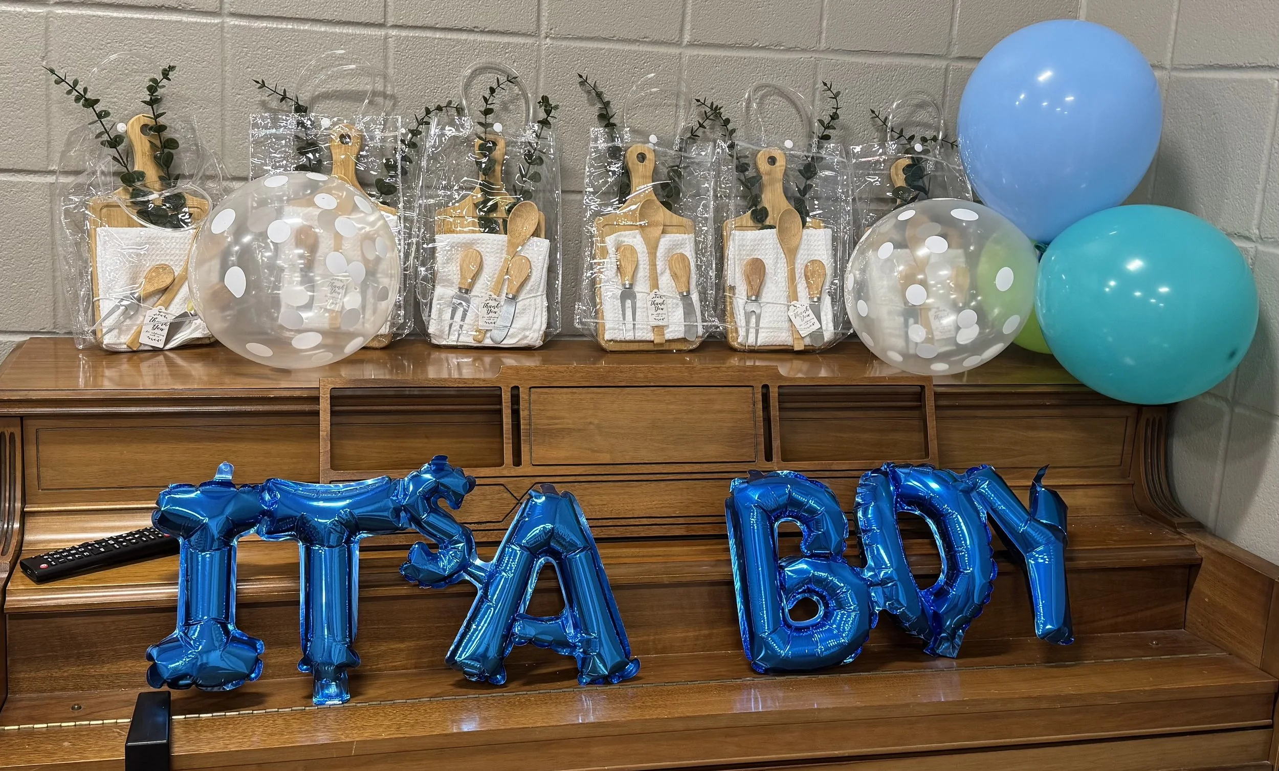Decorative table with blue balloons spelling out 'IT'S A BOY' and gift boxes containing baby-themed items, set against a brick wall.