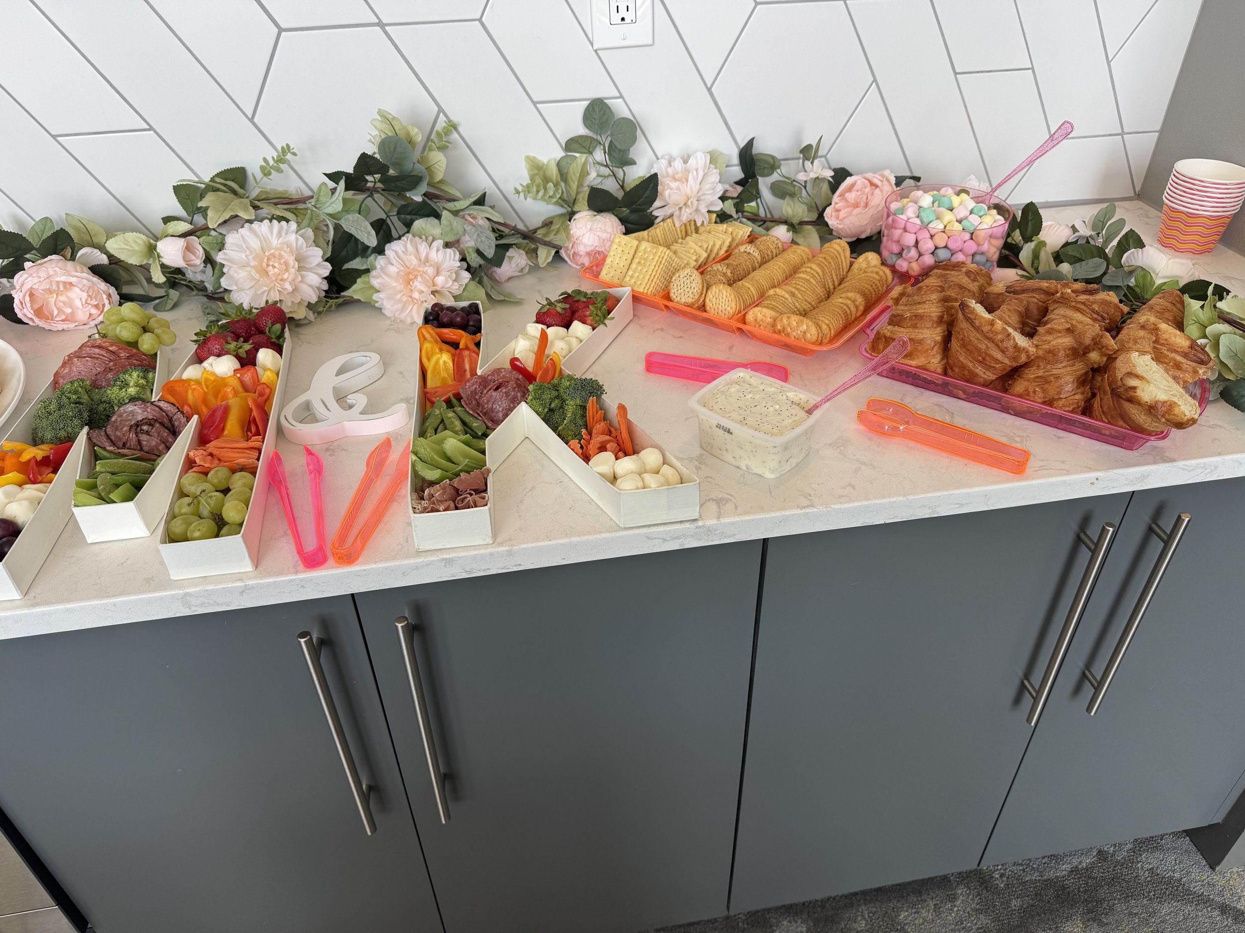 A dessert and snack table with cookies, croissants, marshmallows, fruit, cheese, and vegetables, decorated with a floral garland.