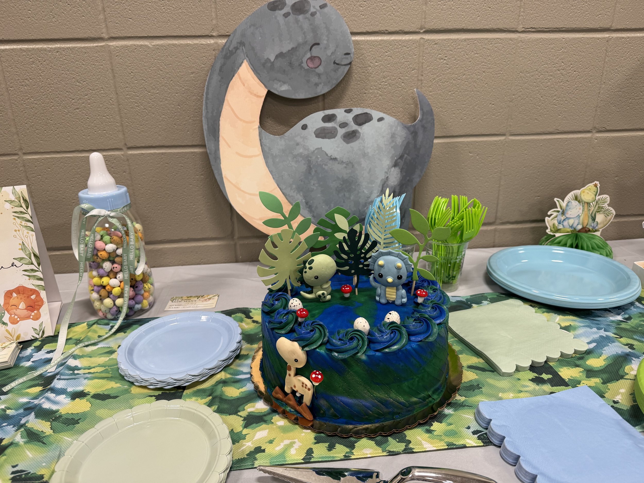 A cake decorated with animal and plant-themed toppers, surrounded by blue and white plates and napkins on a table with a jungle leaf pattern tablecloth. In the background, a moon and bear wall decoration and a bottle of colorful candy olives are visi