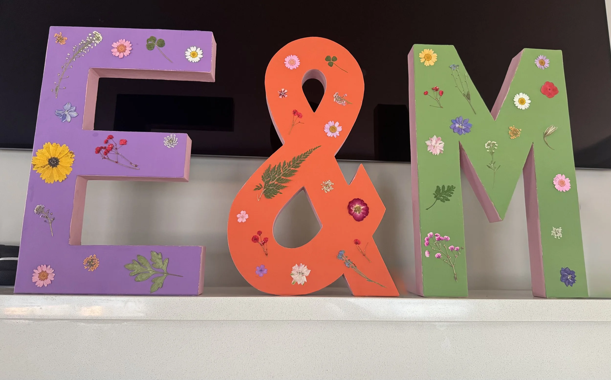 Colorful decorative letters E, &, and M decorated with various pressed flowers and leaves.