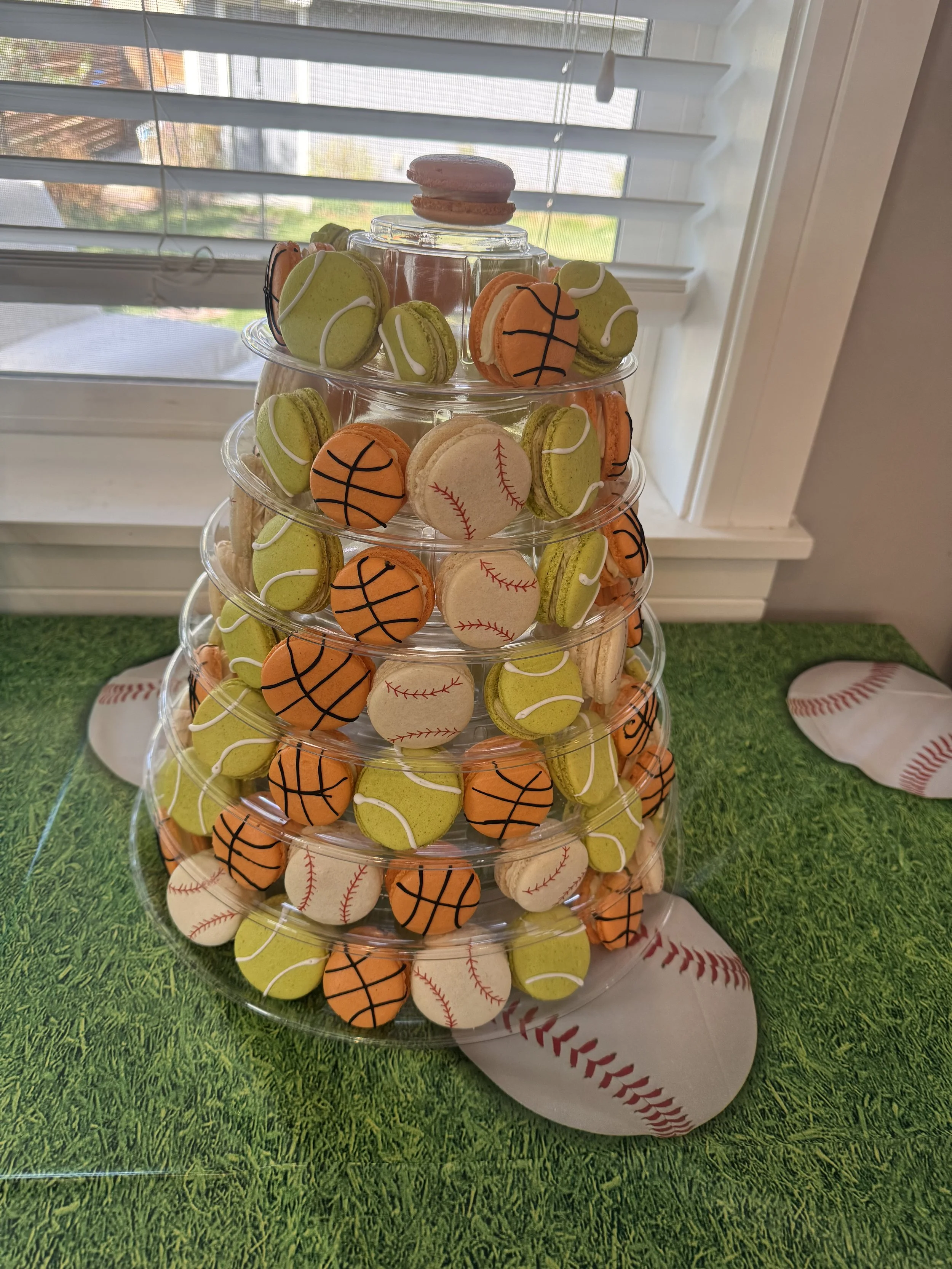Stacked clear plastic containers filled with cookies decorated as baseballs, tennis balls, and basketballs, placed on a table with a baseball field pattern and a grass surface