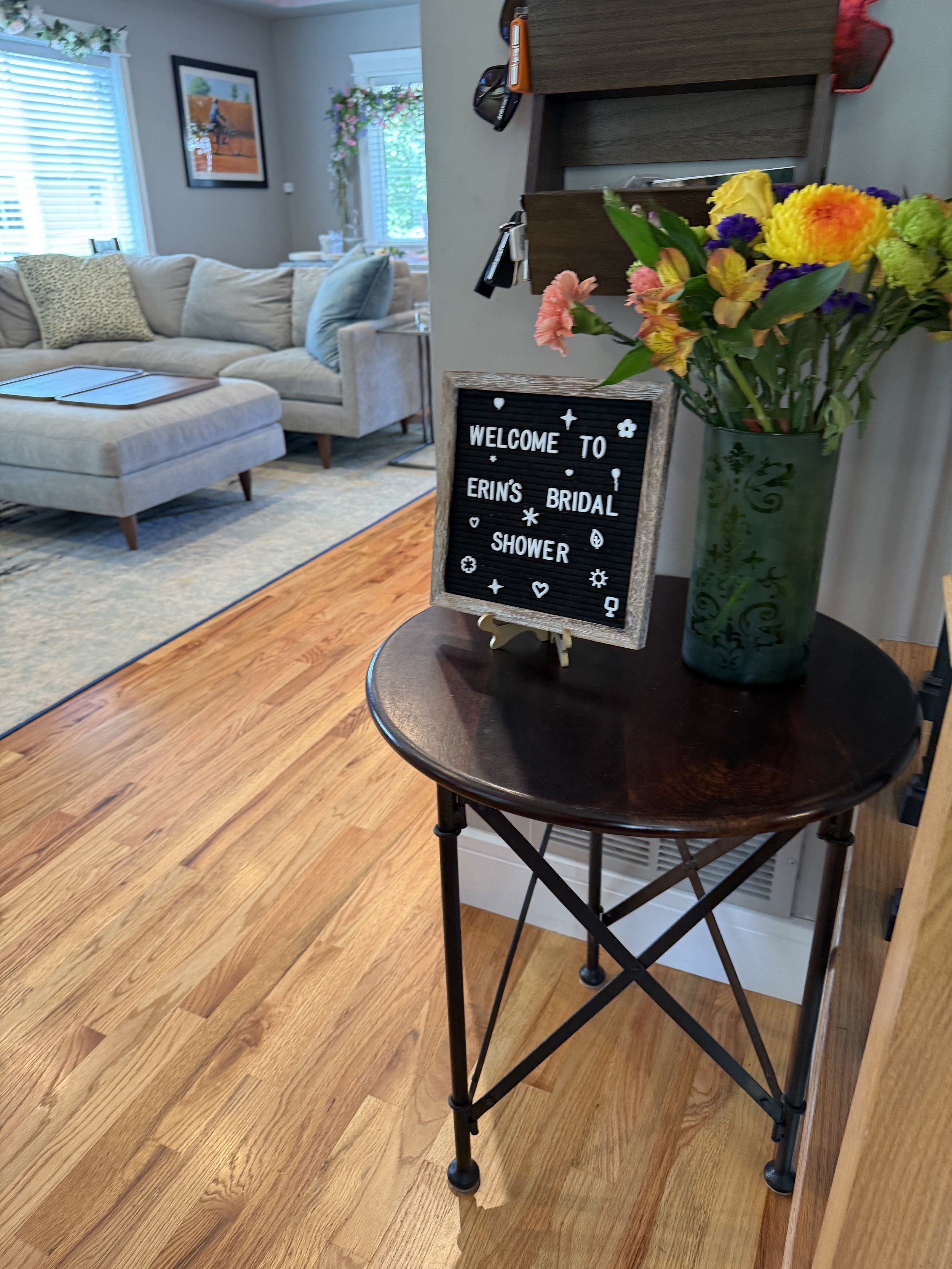 A small round wooden table with a vase of colorful flowers and a welcome sign for Erin's bridal shower in a cozy living room.