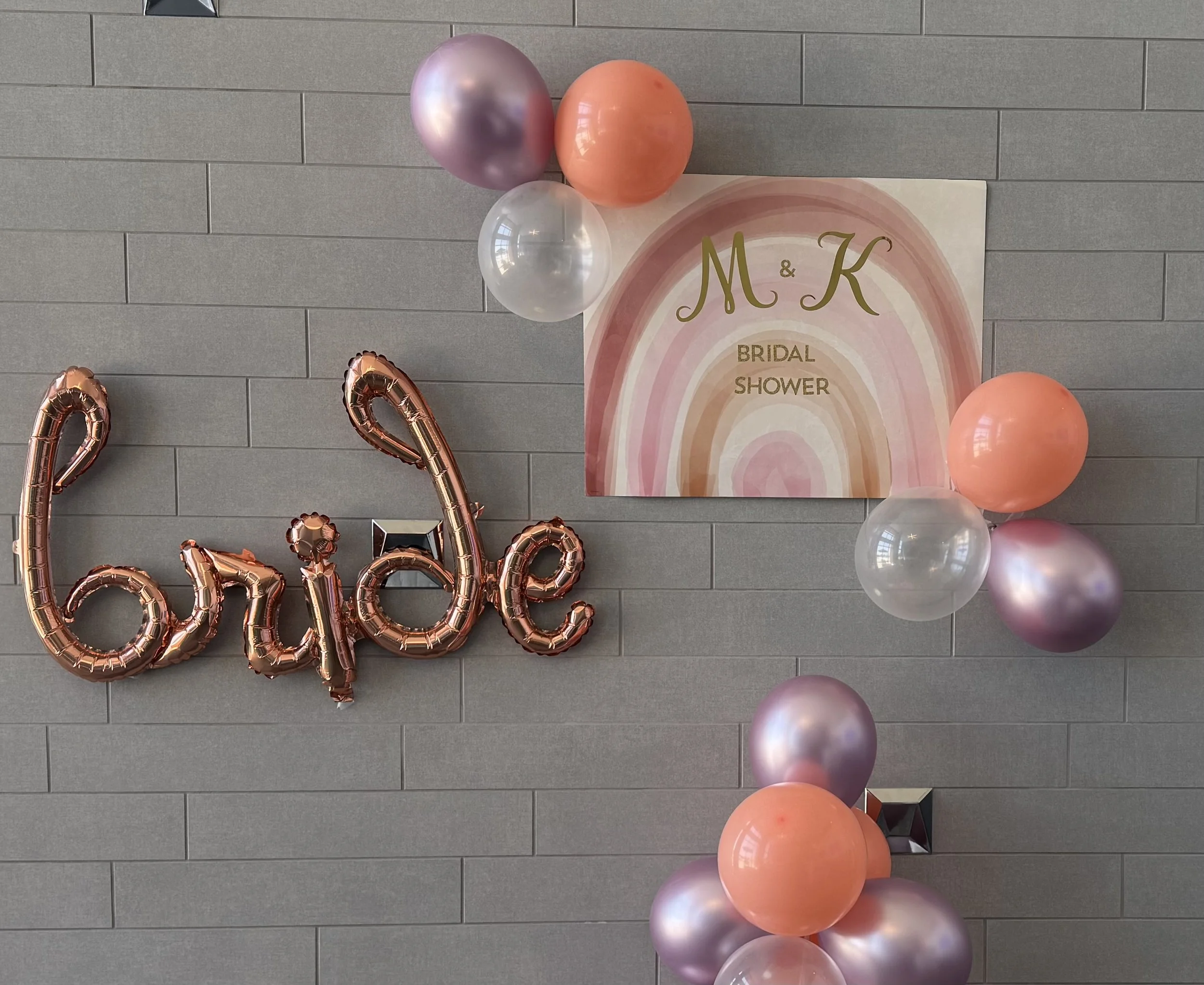 Decorations for a bridal shower featuring a sign with the initials 'M & K' and the words 'Bridal Shower' on a pink and peach watercolor background, accompanied by pink, peach, purple, and white balloons, and a metallic balloon spelling out 'love' in 