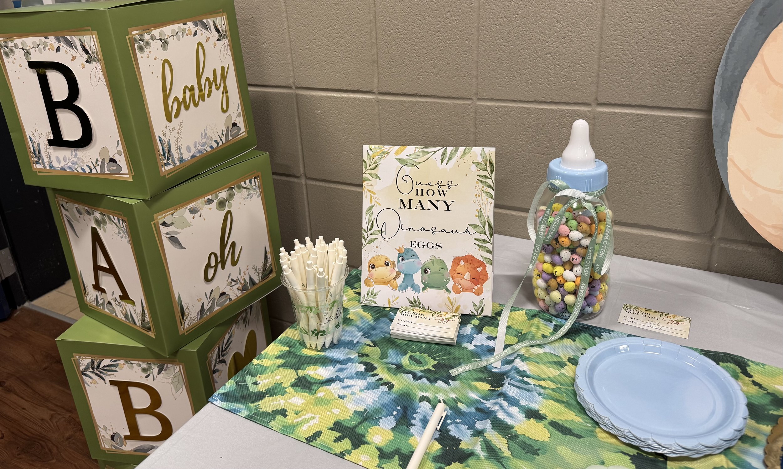 Table decorated for a baby shower with a colorful tablecloth, a stack of blue plates, a jar of colorful candy, and a sign asking how many dinosaur eggs. There are green boxes with letters and words 'baby' and 'oh', a cup with white straws, and a smal