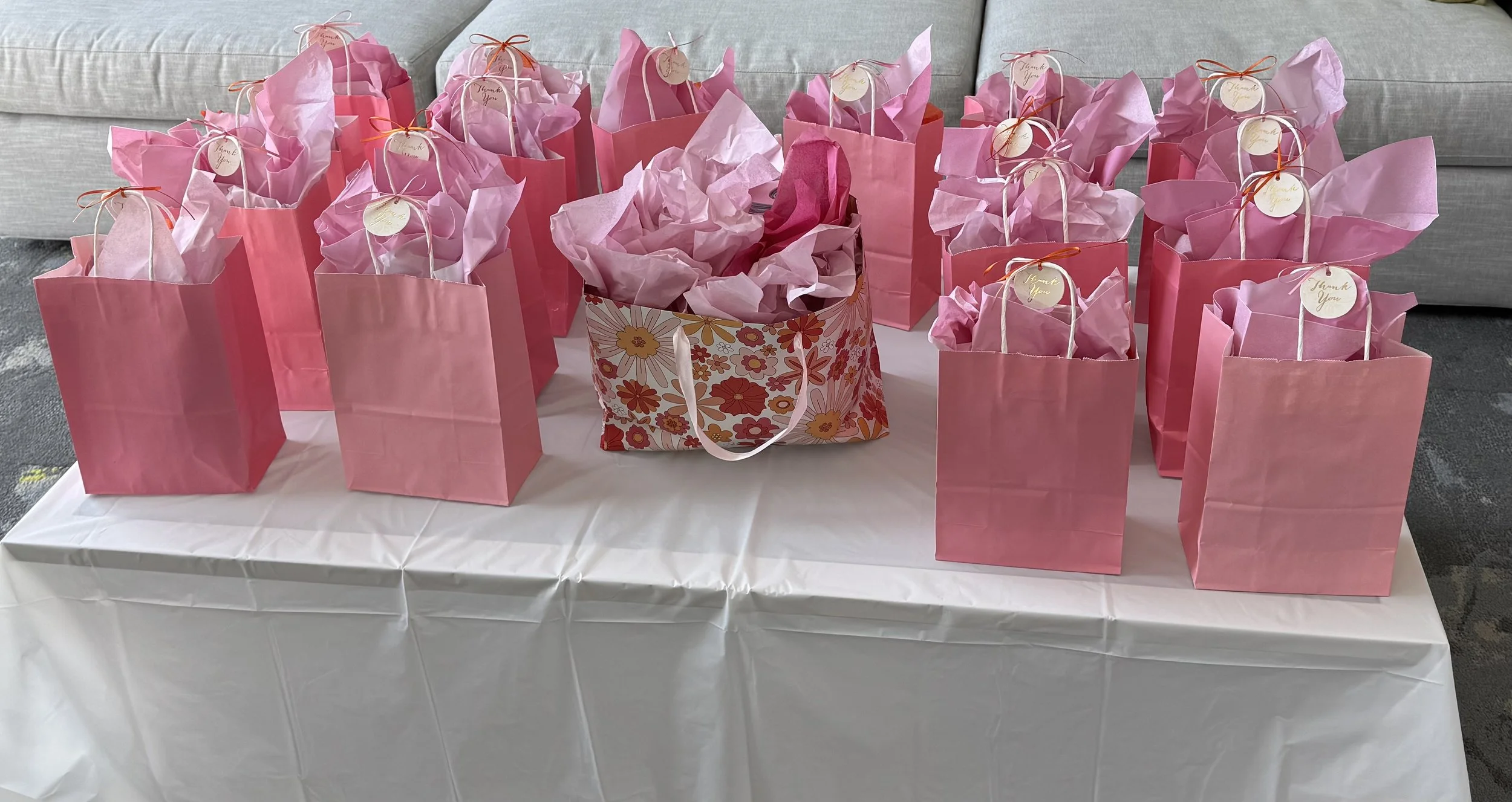 Pink gift bags with tissue paper on a table, some with thank you tags, in front of a gray sofa.
