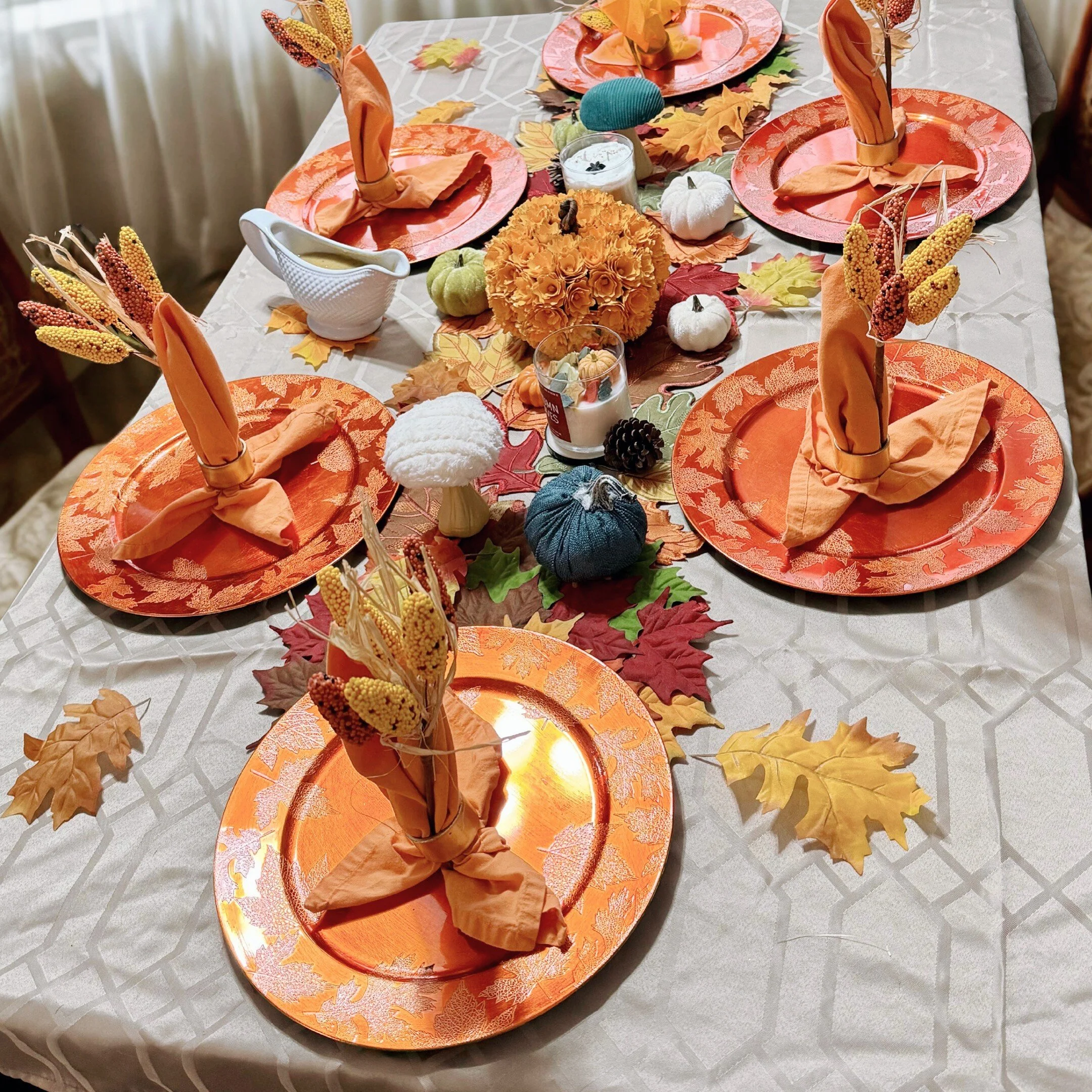 Fall-themed dinner table setting with orange plates, napkins folded in the shape of cones, surrounded by autumn leaves, small pumpkins, and decorative gourds.