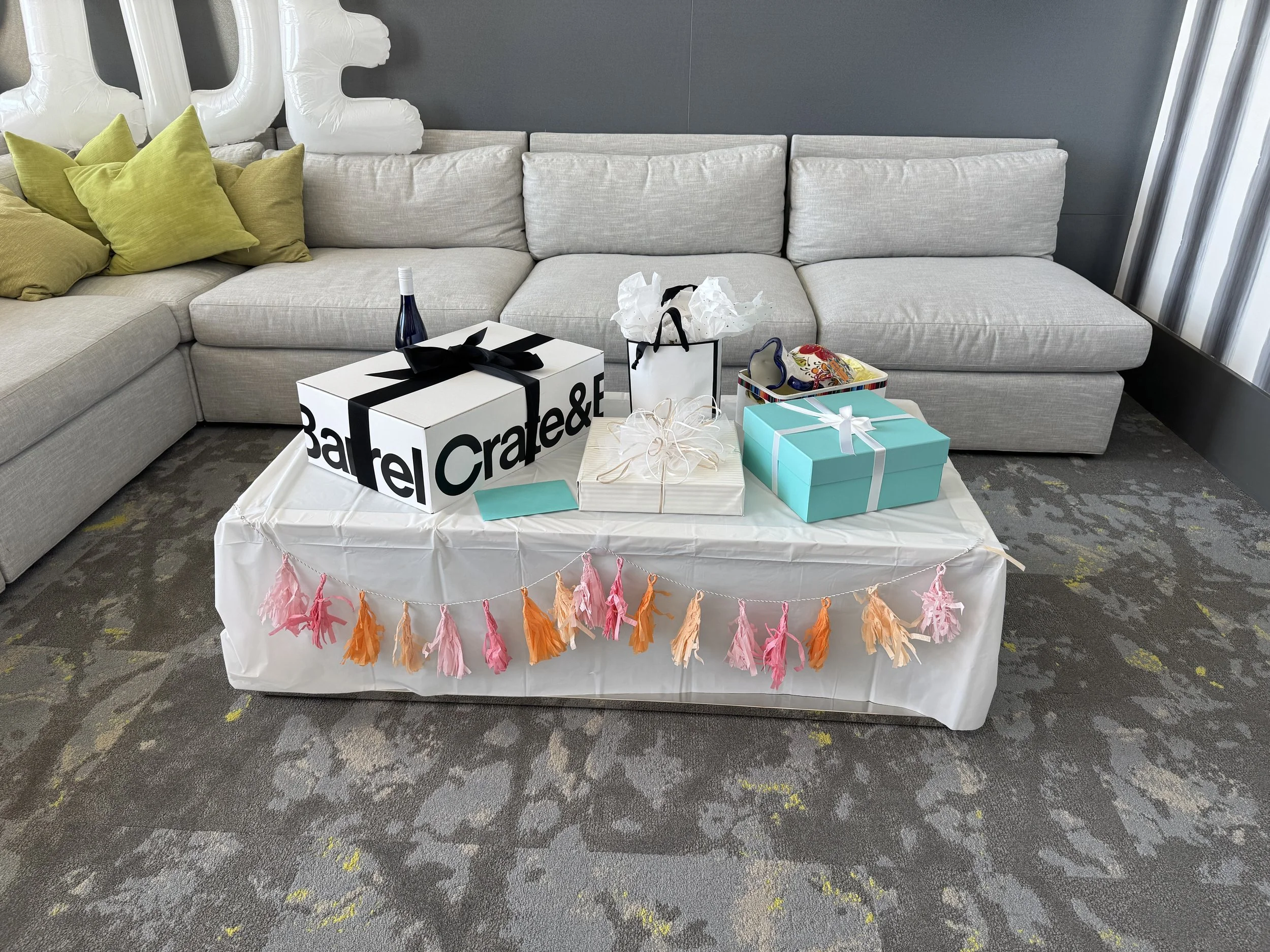 A table with gifts and decorations in front of a light gray sectional sofa with green pillows. The table has a pink, orange, and peach tissue paper tassel garland hanging from it.