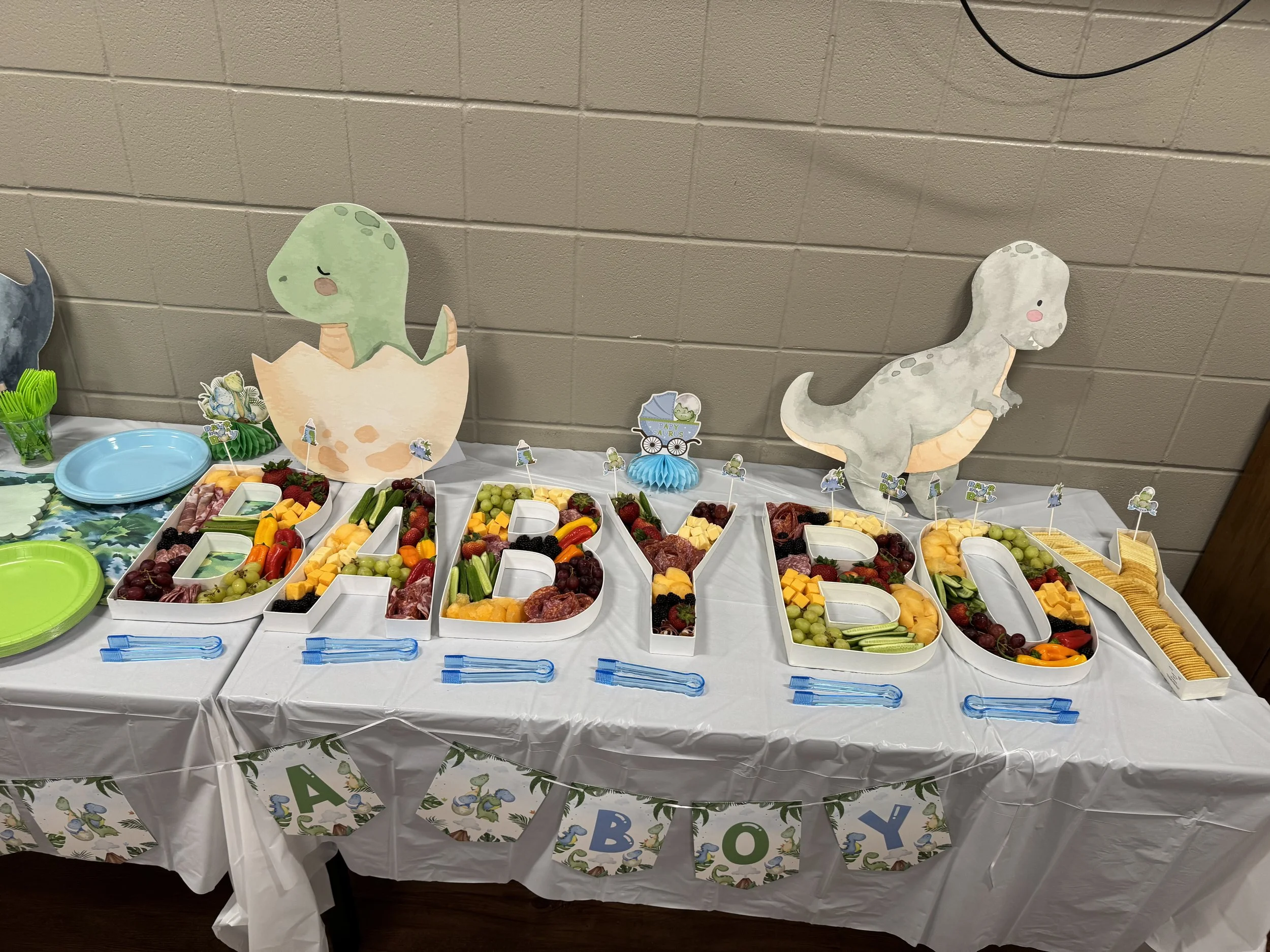 A baby shower table decorated with a dinosaur theme featuring large decorated letters spelling 'BABY' filled with fruits and snacks, two dinosaur-themed cardboard cutouts, small flags with dinosaurs, and colorful plates and utensils.