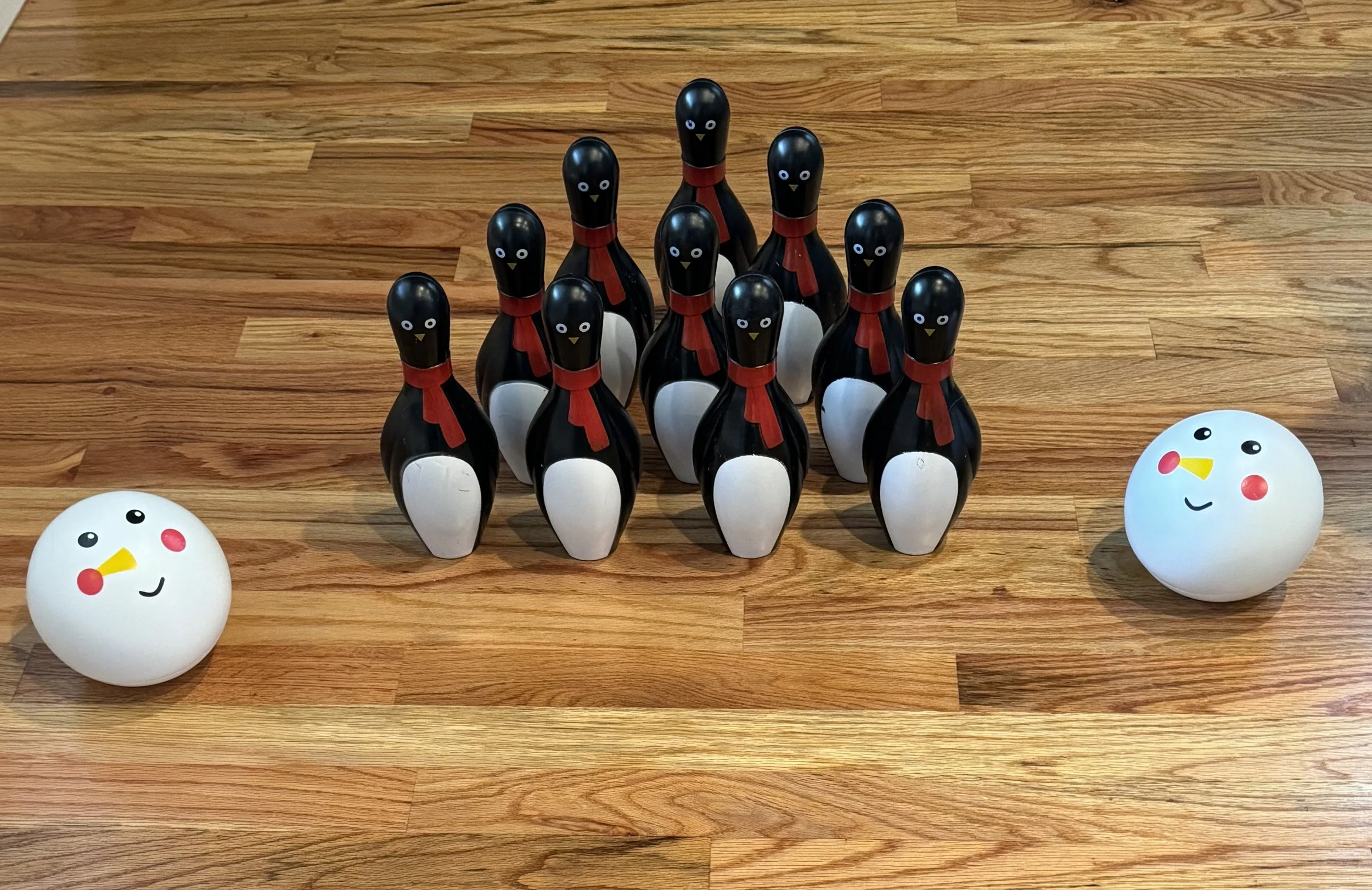 A group of nine penguin-shaped bowling pins, each with a black body, white belly, red scarf, and painted eyes and beak, arranged in formation on a wooden floor. Two large, round, white, smiley face balls with blush on cheeks and beak are positioned o