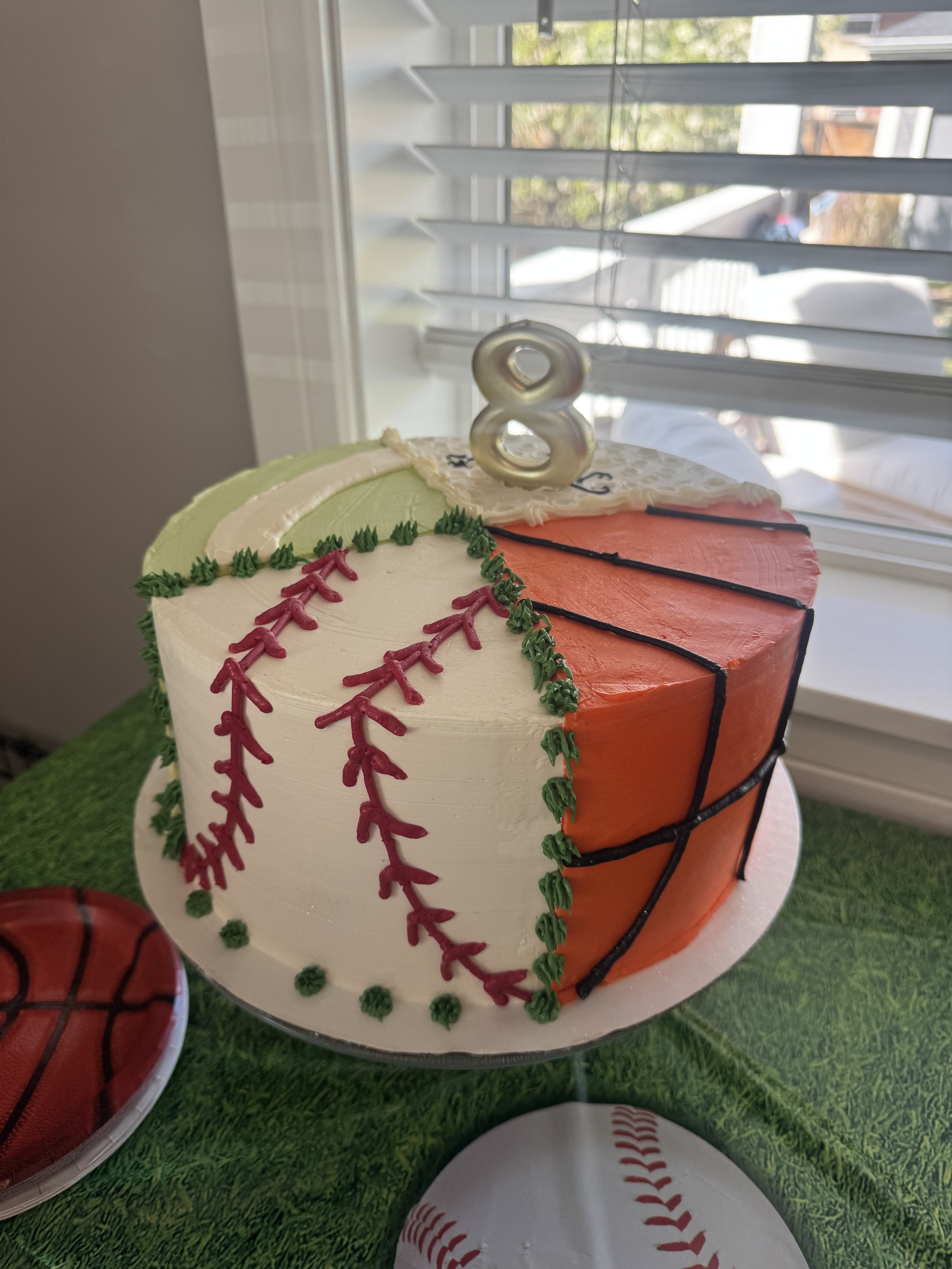 A baseball-themed cake decorated with white, orange, and white frosting with red stitches and green piping, topped with a silver number 8 candle.