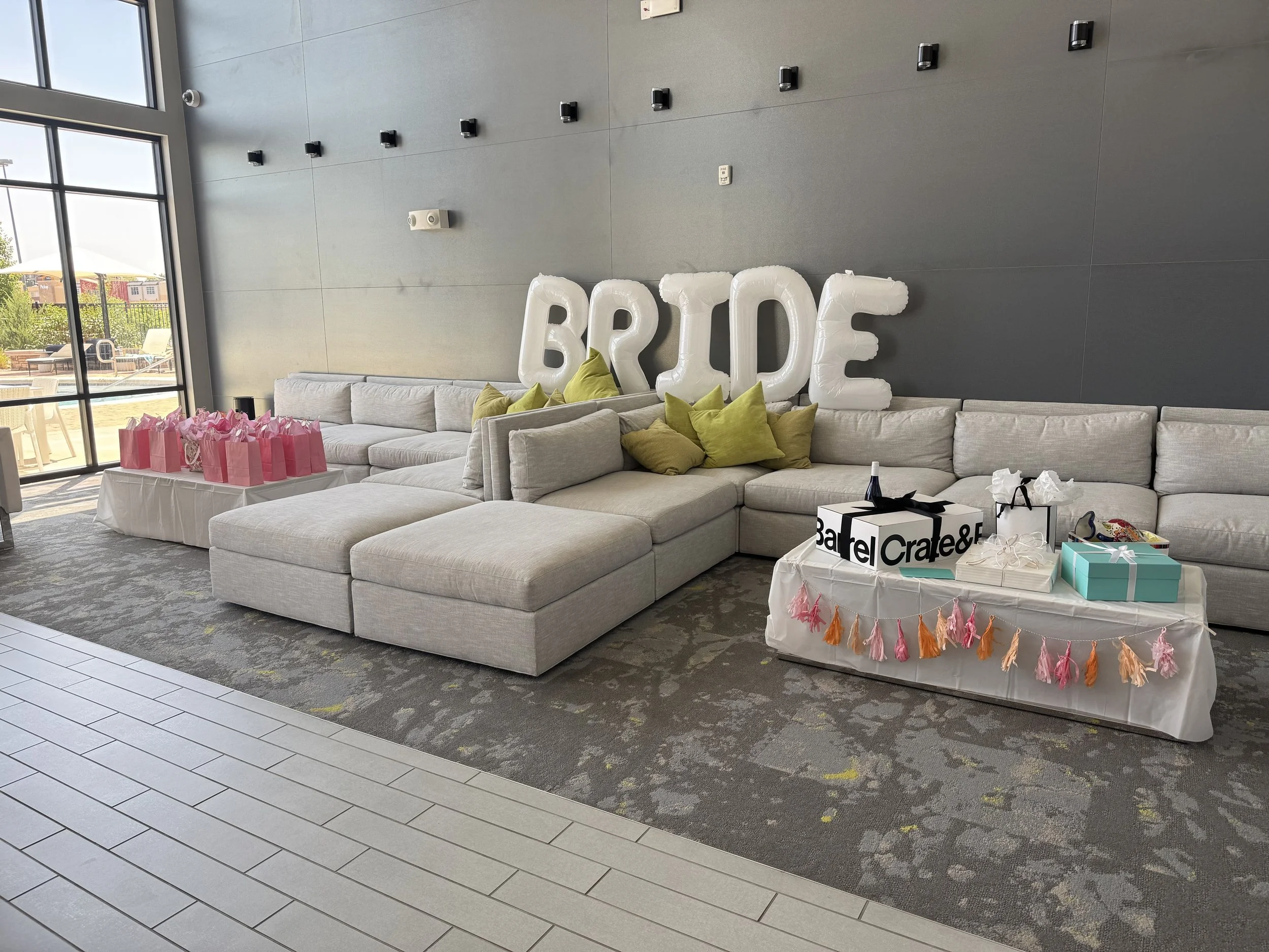 Decorated lounge area with large white balloons spelling 'BRIDE' on the gray wall, a long gray sectional sofa with green and beige pillows, tables with pink gift bags, and a table with wrapped gifts and decorative tassels for a bridal celebration.