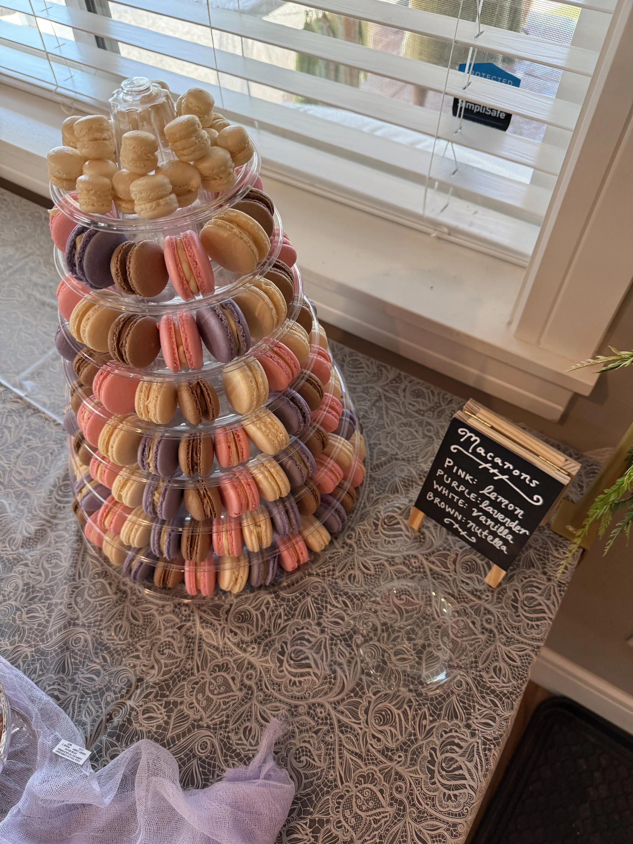 A display of colorful macarons stacked in a clear plastic cone, next to a small blackboard sign listing flavors such as pink lemon, purple lavender, white vanilla, and brown nutella, on a lace-patterned table near a window.