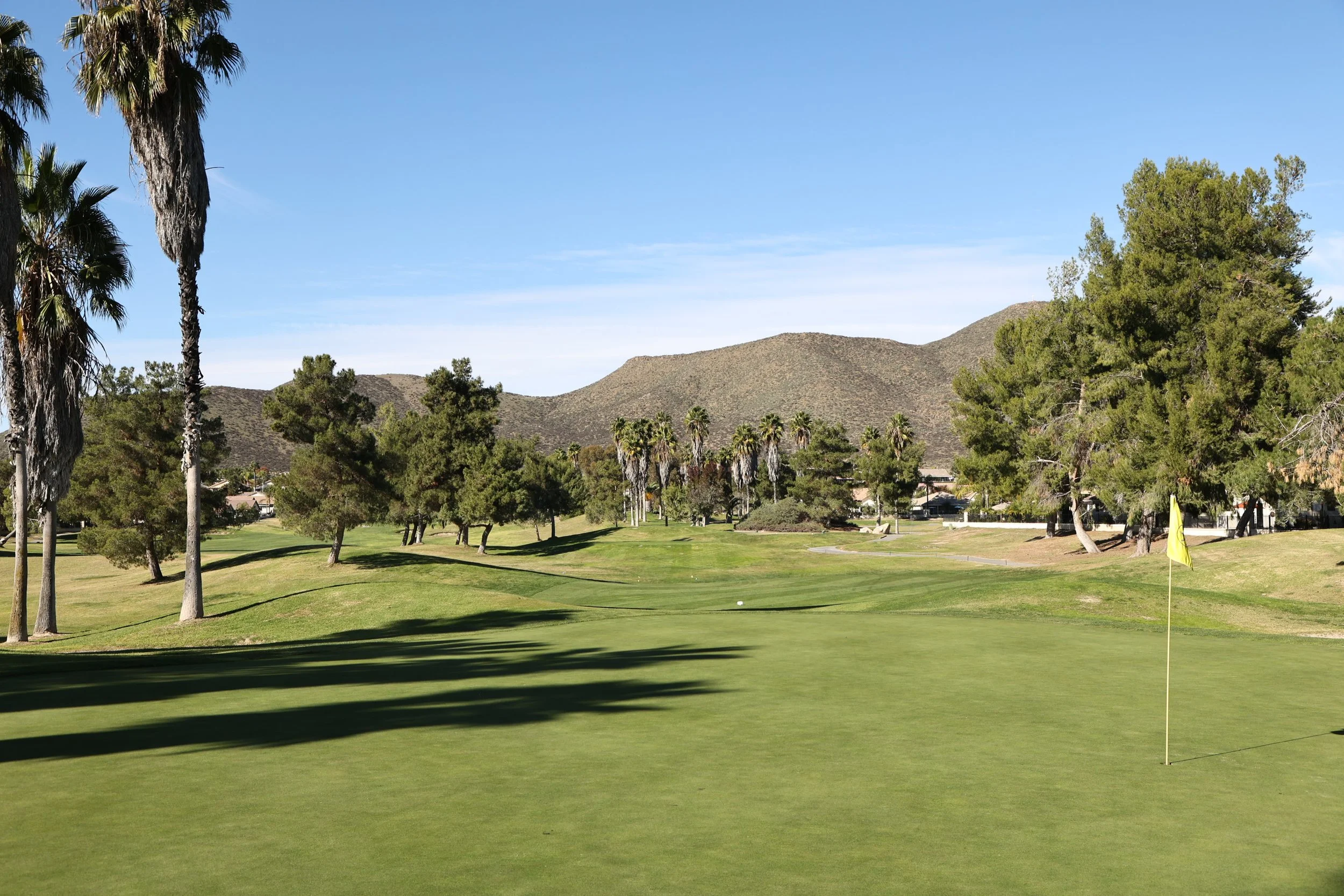 A golf course with green grass, trees, and mountains in the background under a blue sky.