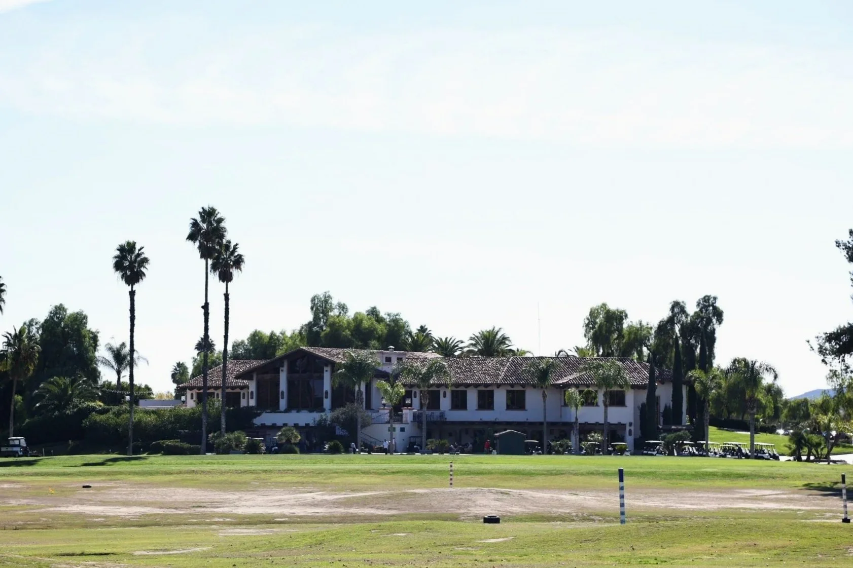 Menifee Lakes Country Club Clubhouse with a tiled roof, surrounded by tall palm trees, on a well-manicured golf course with green grass, golf clubs and carts nearby, under a clear blue sky.