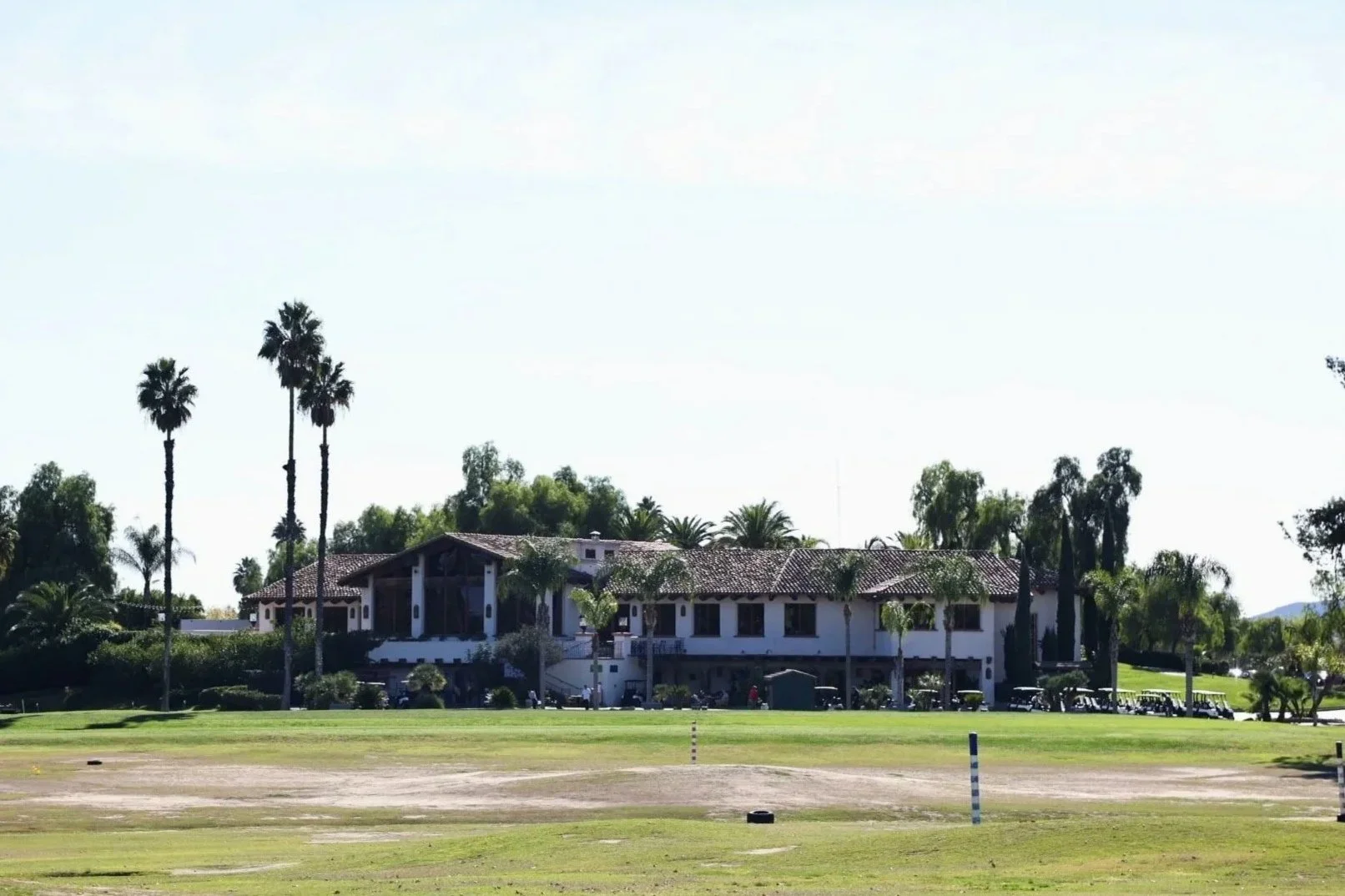 Menifee Lakes Country Club Clubhouse with a tiled roof, surrounded by tall palm trees, on a well-manicured golf course with green grass, golf clubs and carts nearby, under a clear blue sky.