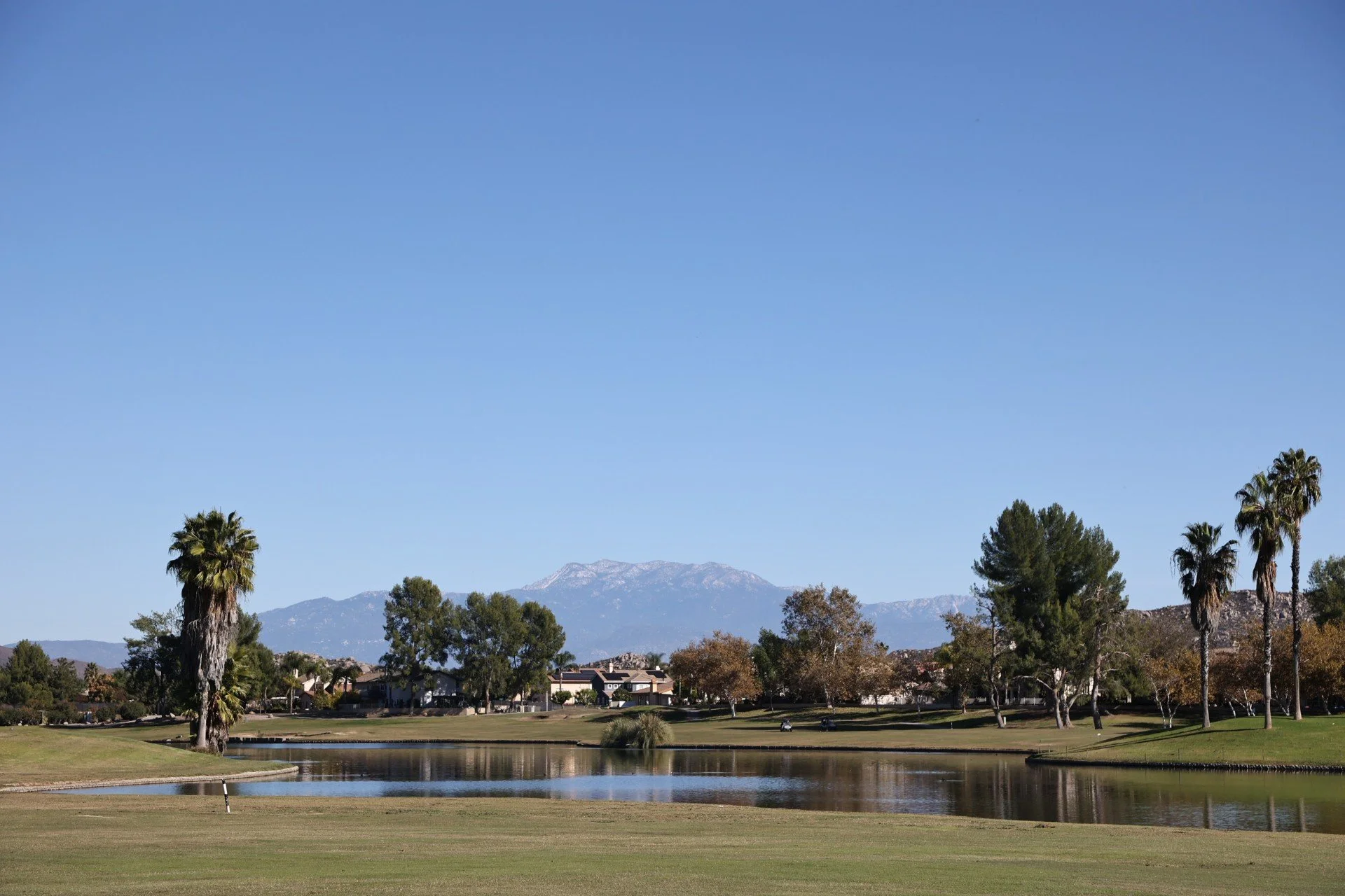 A peaceful golf course with a pond, tall palm trees, and mountains in the distance under a clear blue sky.