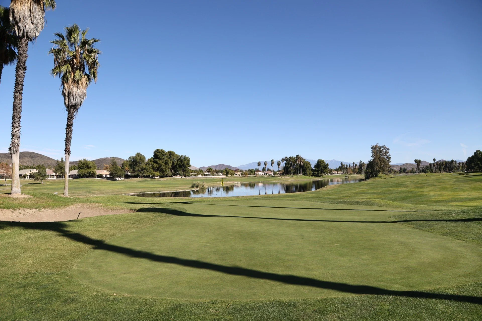 View of a golf course with green grass, palm trees, a small water hazard, and distant mountains under a clear blue sky.