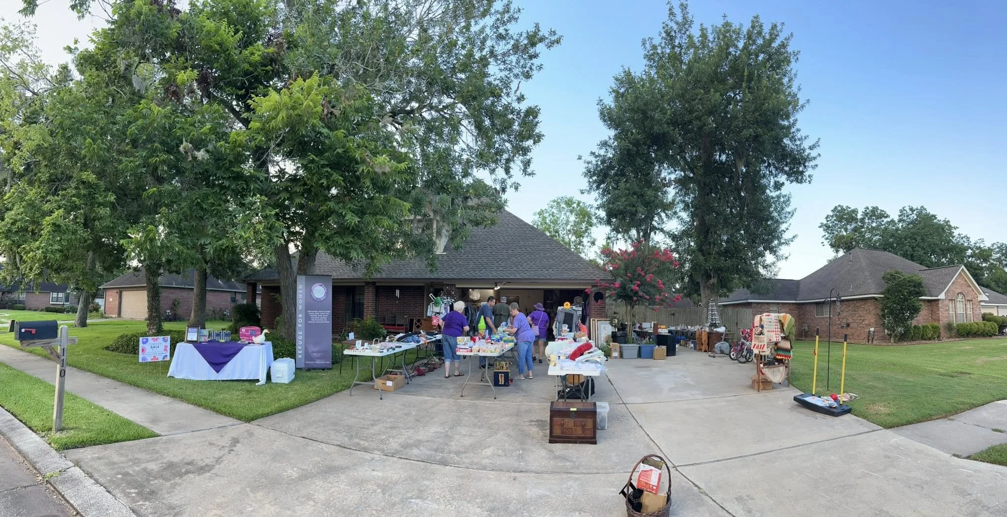 A garage sale set up in a driveway with tables and boxes of various items, a few people shopping, trees, and houses in a suburban neighborhood on a clear day.