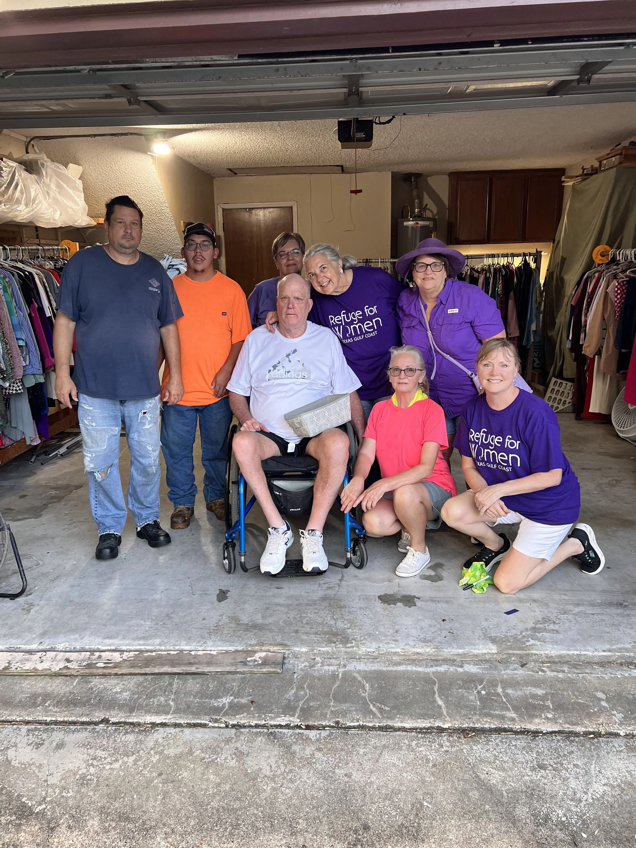 A group of eight people, including an elderly man in a wheelchair, gathered in a garage with clothes racks and a closed garage door. Some wear matching 'Refuge for Women' shirts.