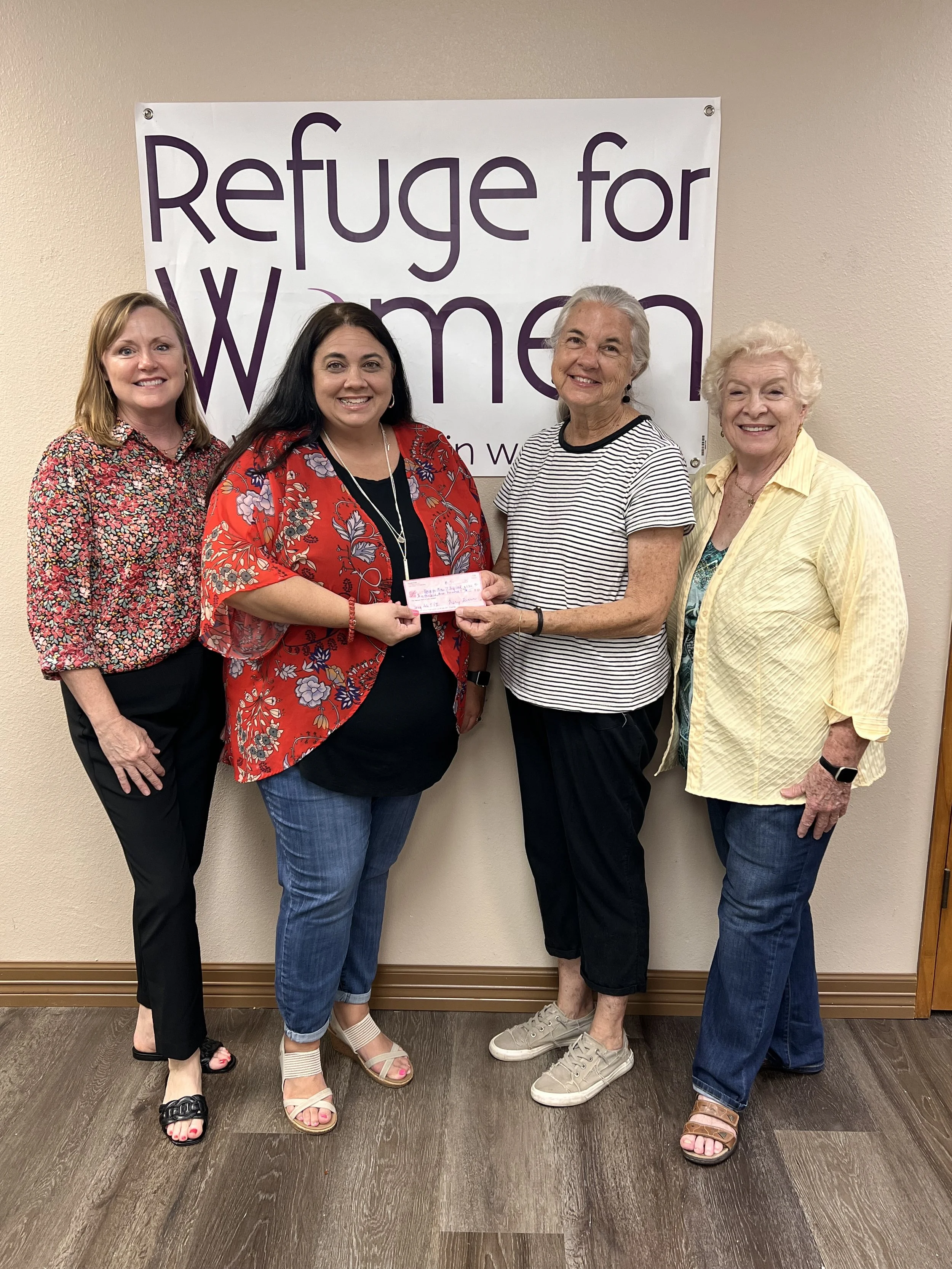 Four women standing in front of a banner that reads 'Refuge for Women,' with one woman handing a check to another.